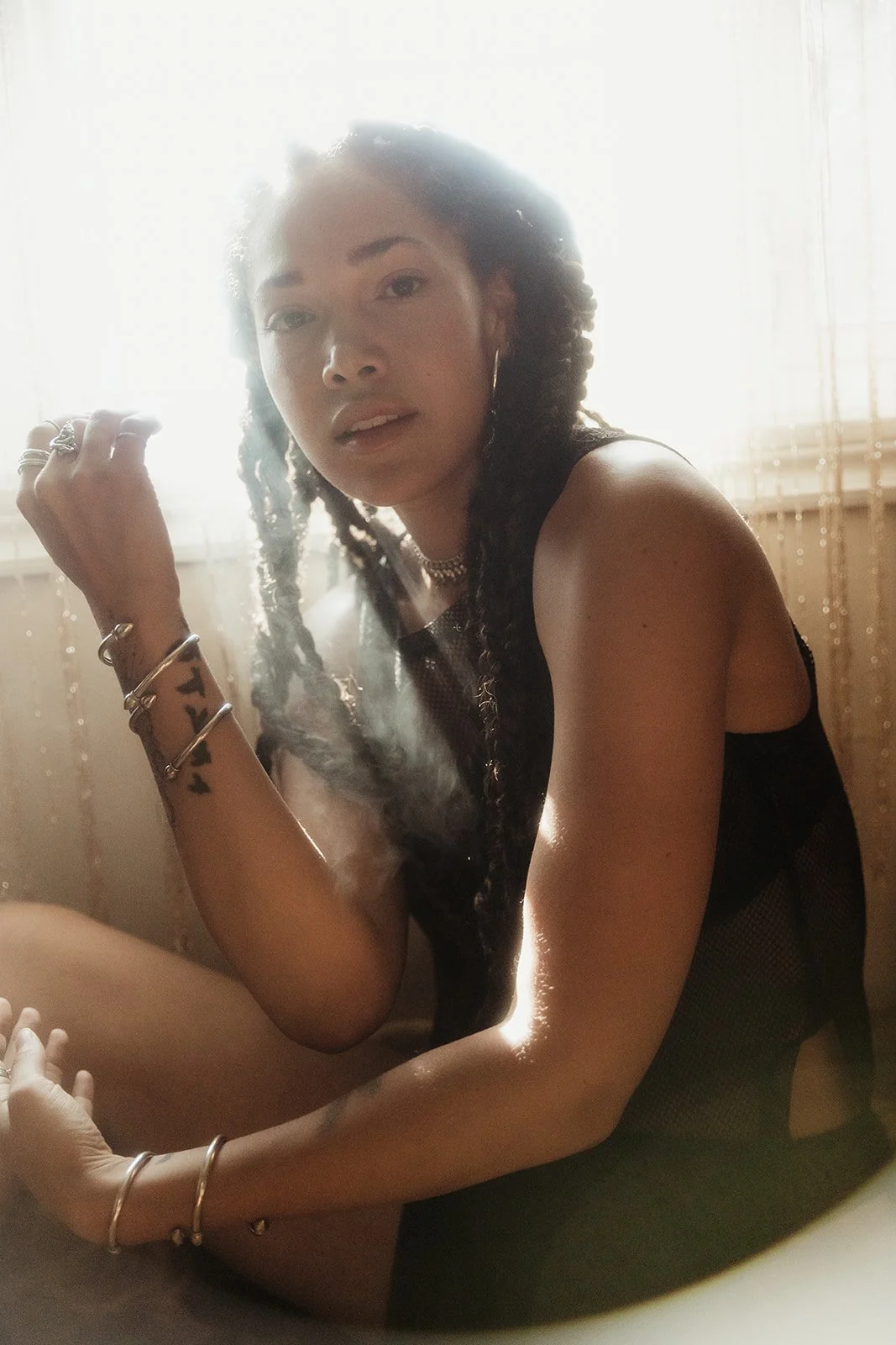 Person with braided hair sitting in soft lighting, wearing jewelry and a sleeveless top.