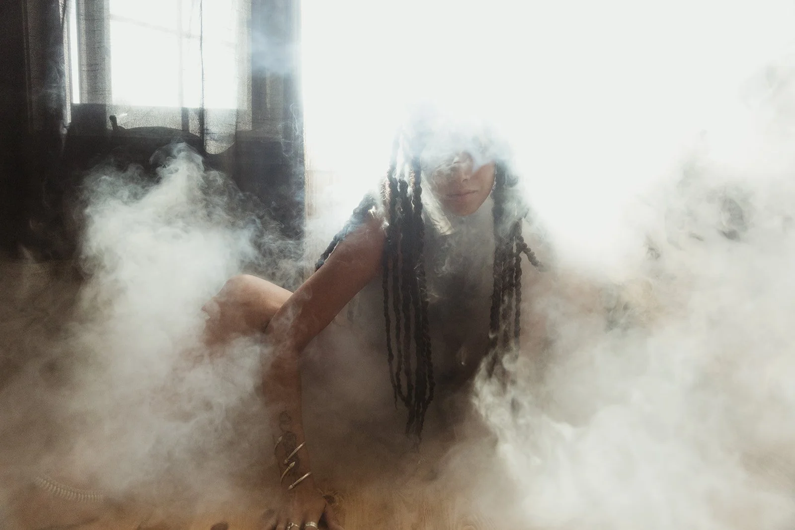 Person with long braids shrouded in smoke, crouching on the floor in a dimly lit room.