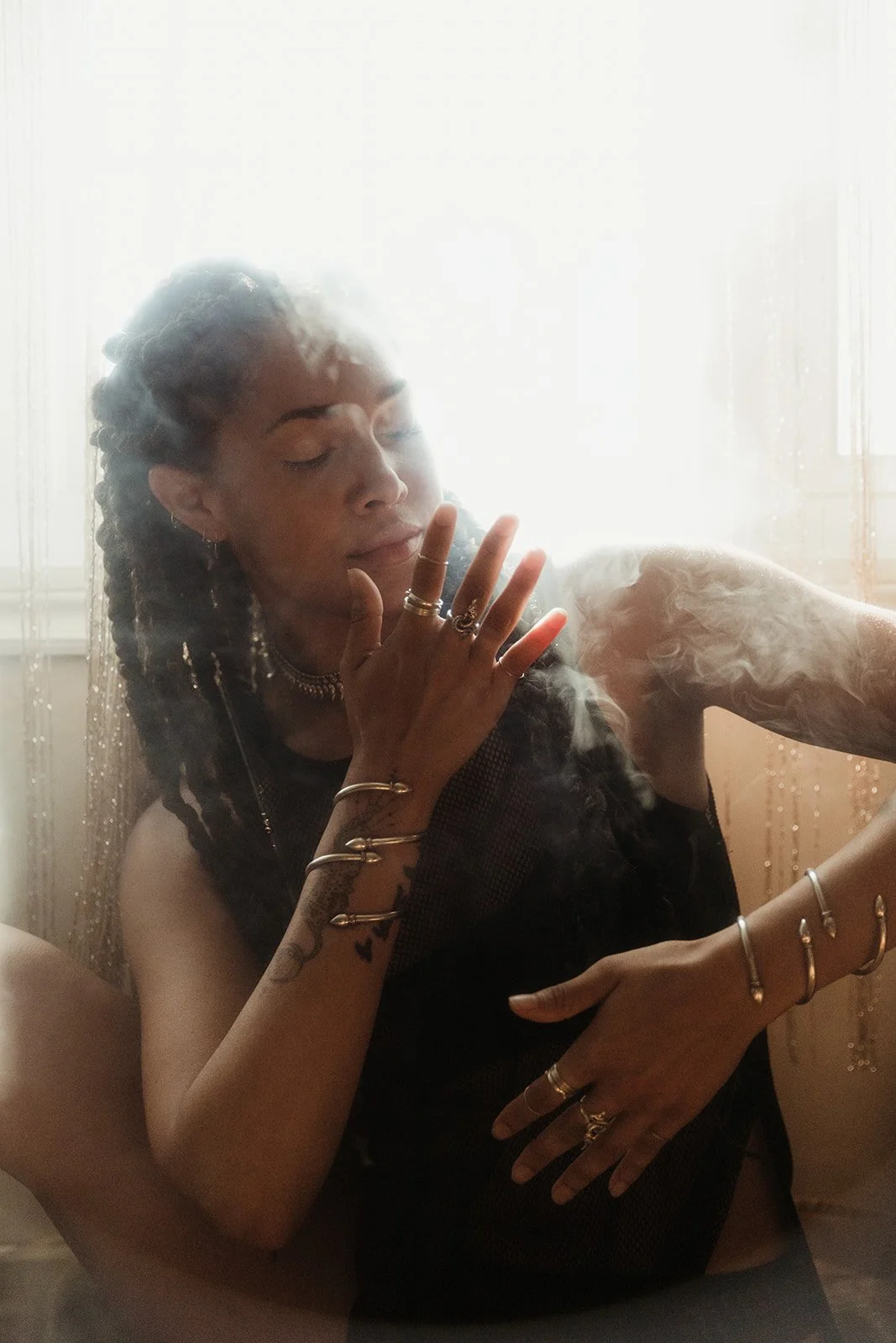 Person with braided hair, wearing multiple rings and bracelets, surrounded by smoke, sitting in soft lighting.
