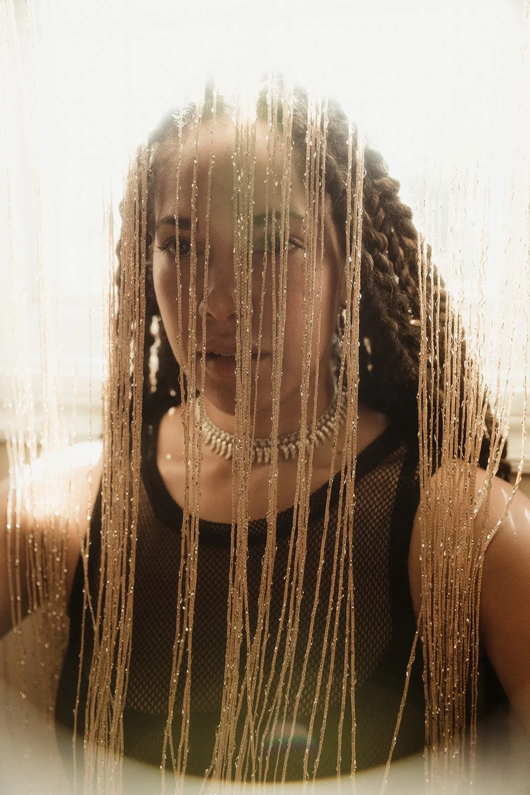 Woman with braided hair behind beaded curtain, wearing black mesh top and necklace.