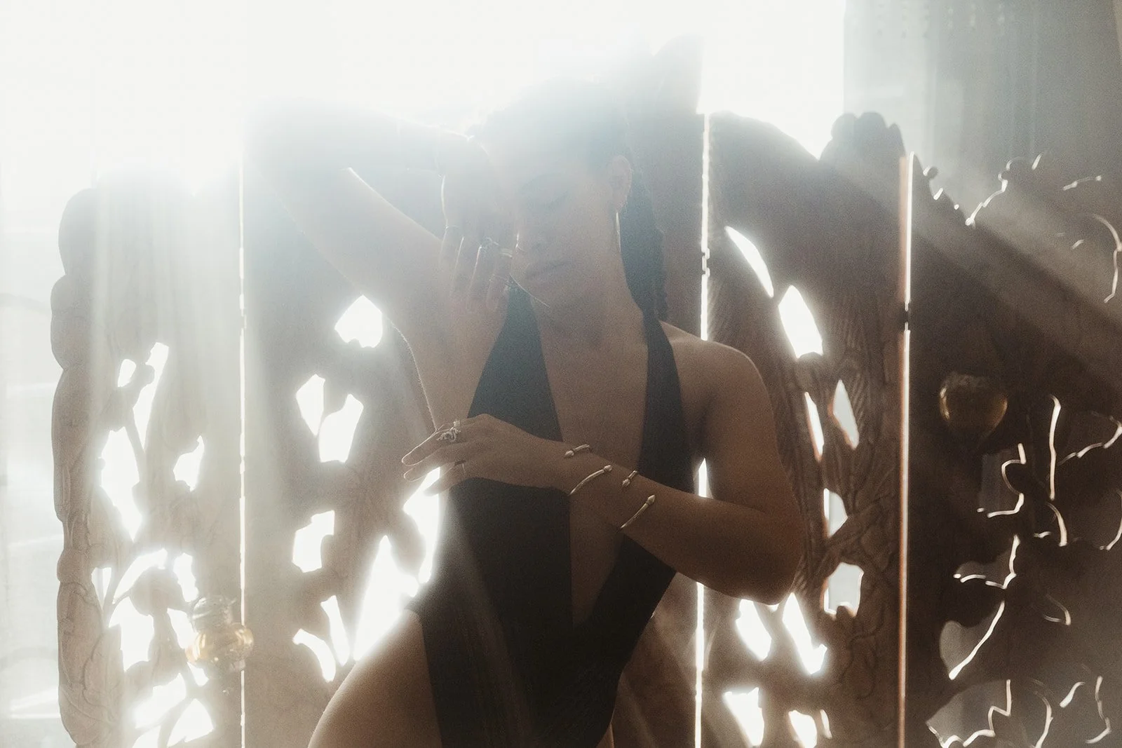 Person in black outfit posing against backlit ornate wooden screen with sunlight streaming through.