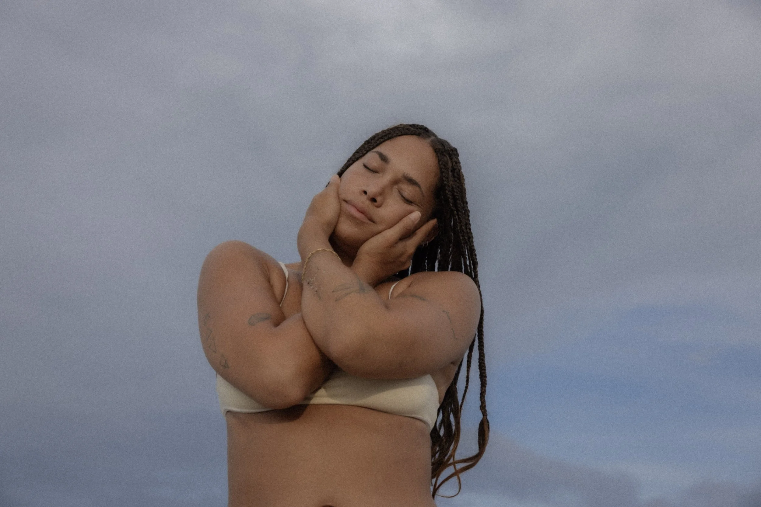 Person with braided hair wearing white bikini top, eyes closed, and hands gently on face in relaxed pose against cloudy sky.