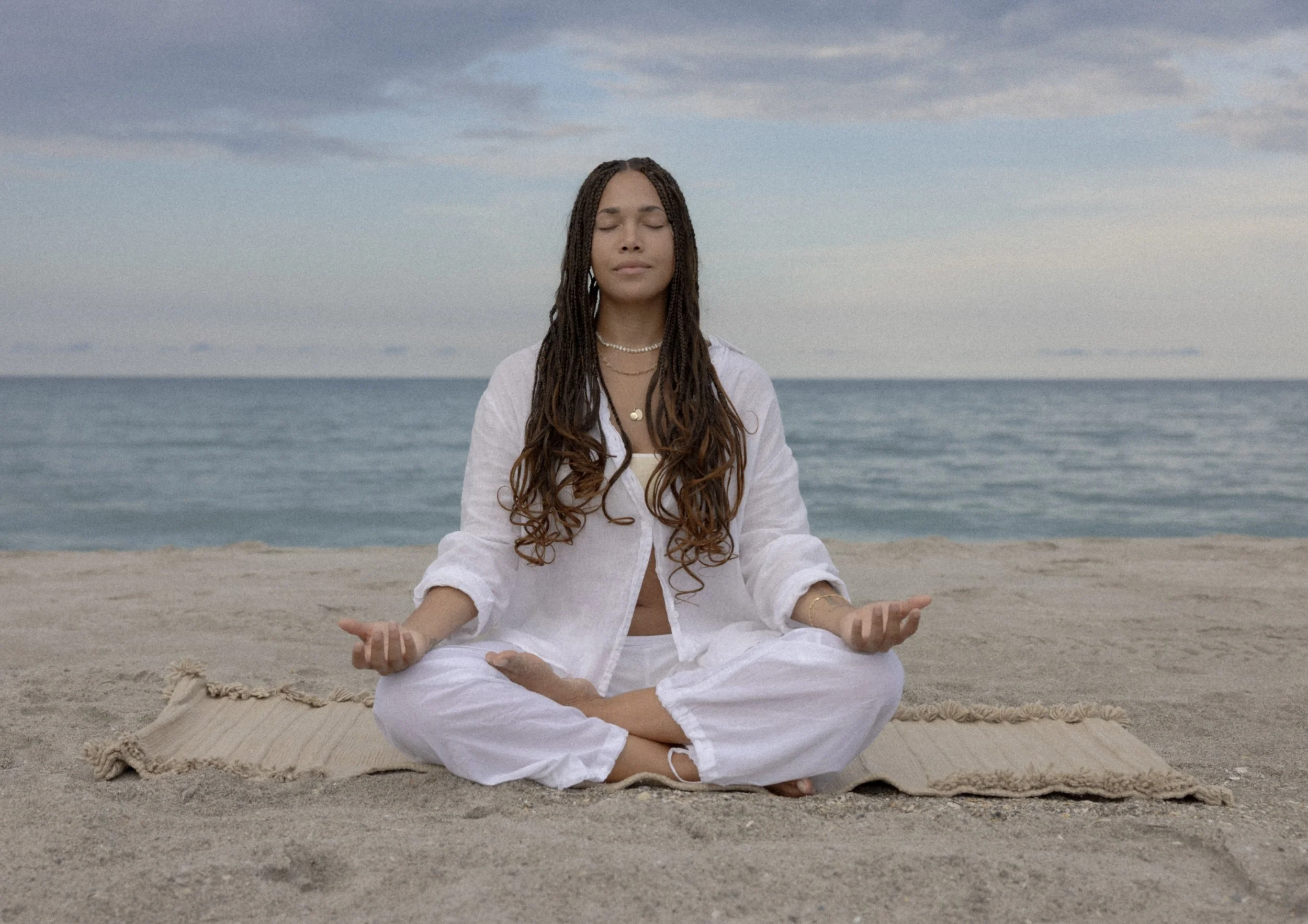 A person meditating on a beach wearing a white outfit, eyes closed, sitting cross-legged on a mat, with the ocean in the background.