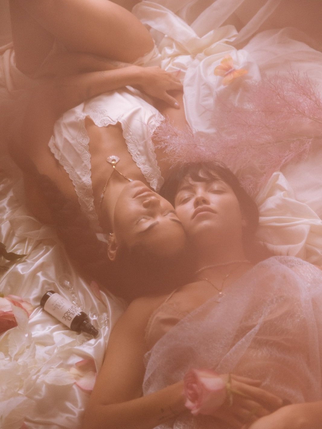 Two individuals lying on a bed surrounded by pink and white fabric, holding flowers. A bottle is beside them.