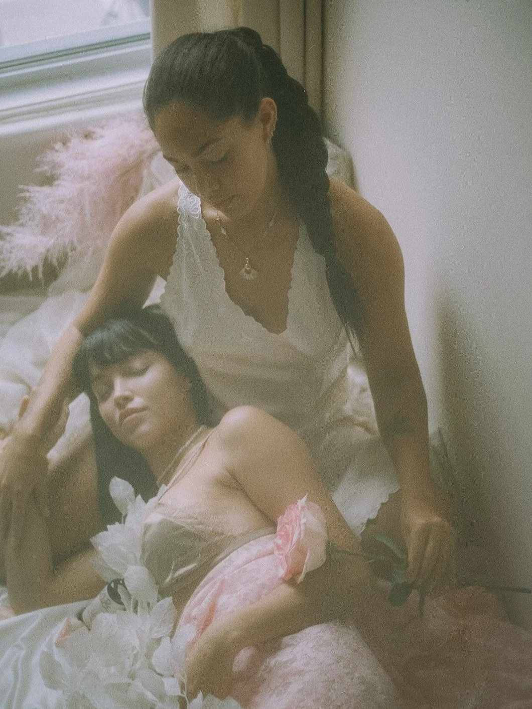 Two people in soft pastel clothing, sitting and lying on a bed next to each other, with one holding a rose and surrounded by flowers, in a softly lit room.