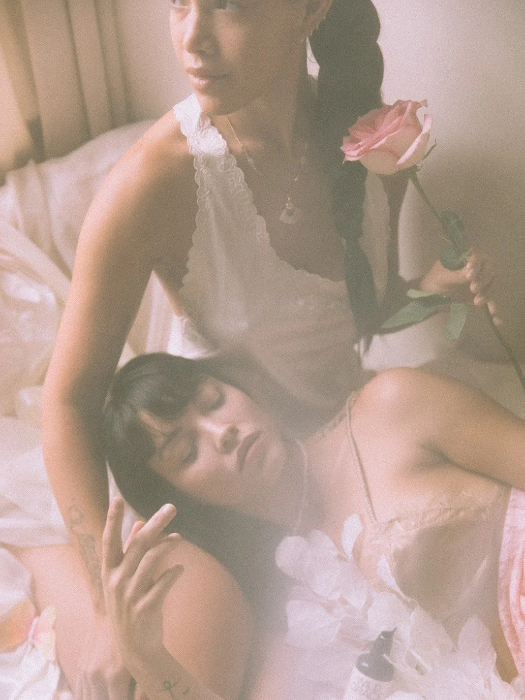 Two women in delicate clothing, one holding a pink rose while the other rests her head on the other's lap, creating a serene, intimate atmosphere.