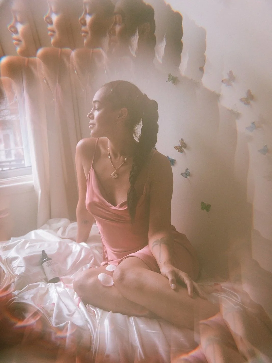 Woman in pink dress sitting on bed with multiple exposure effect, surrounded by butterfly wall decor and petals, bottle of oil on bed.