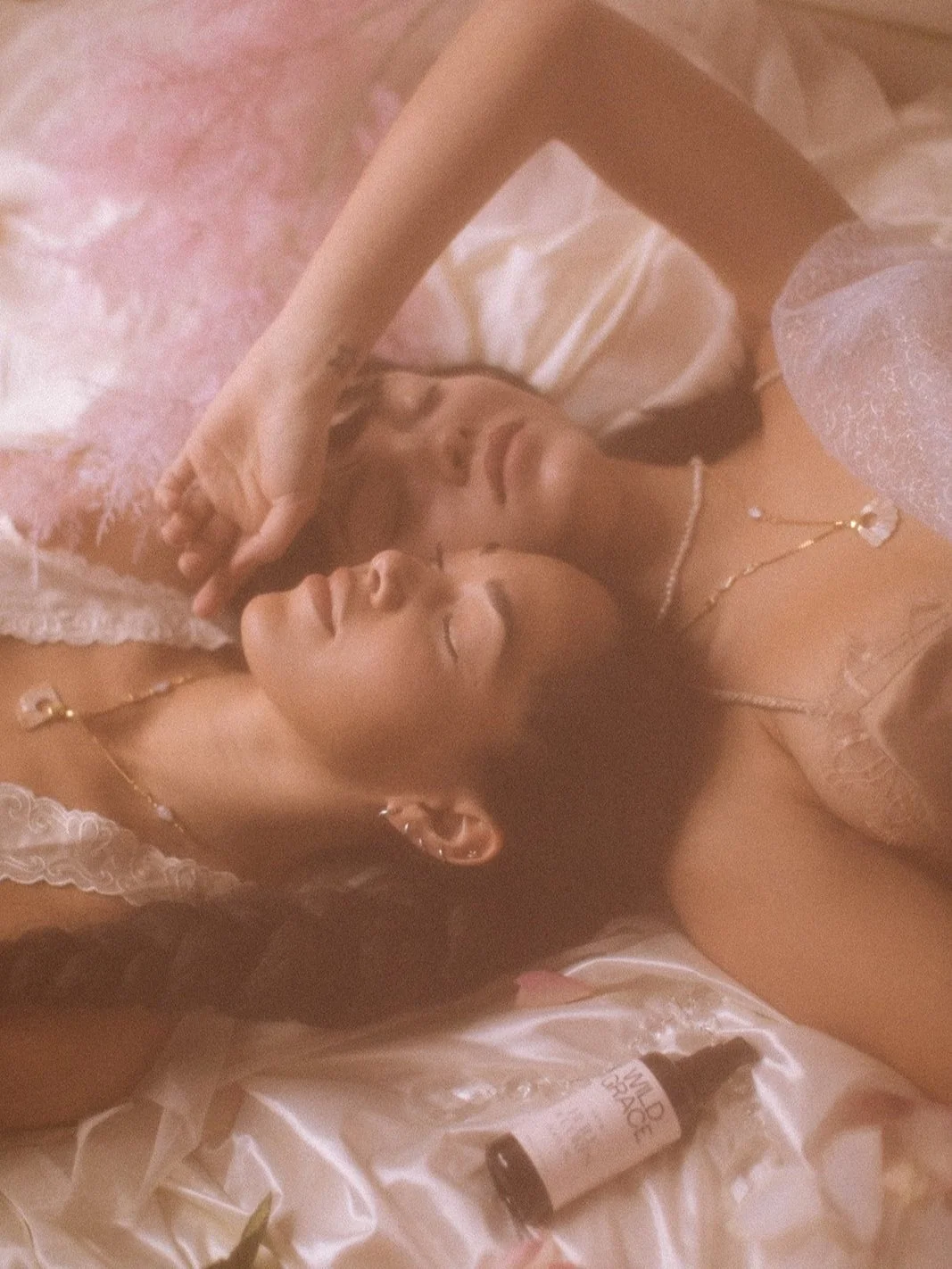 Two women lying on satin sheets with a bottle labeled "Wild Sage" nearby, wearing lace garments, surrounded by a soft, dreamy atmosphere.