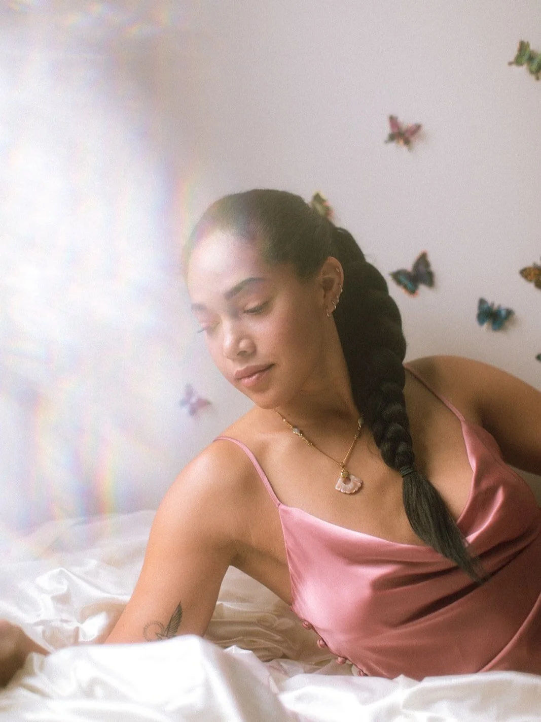 Woman in pink dress with braided hair sitting on a bed with butterfly decorations on the wall.