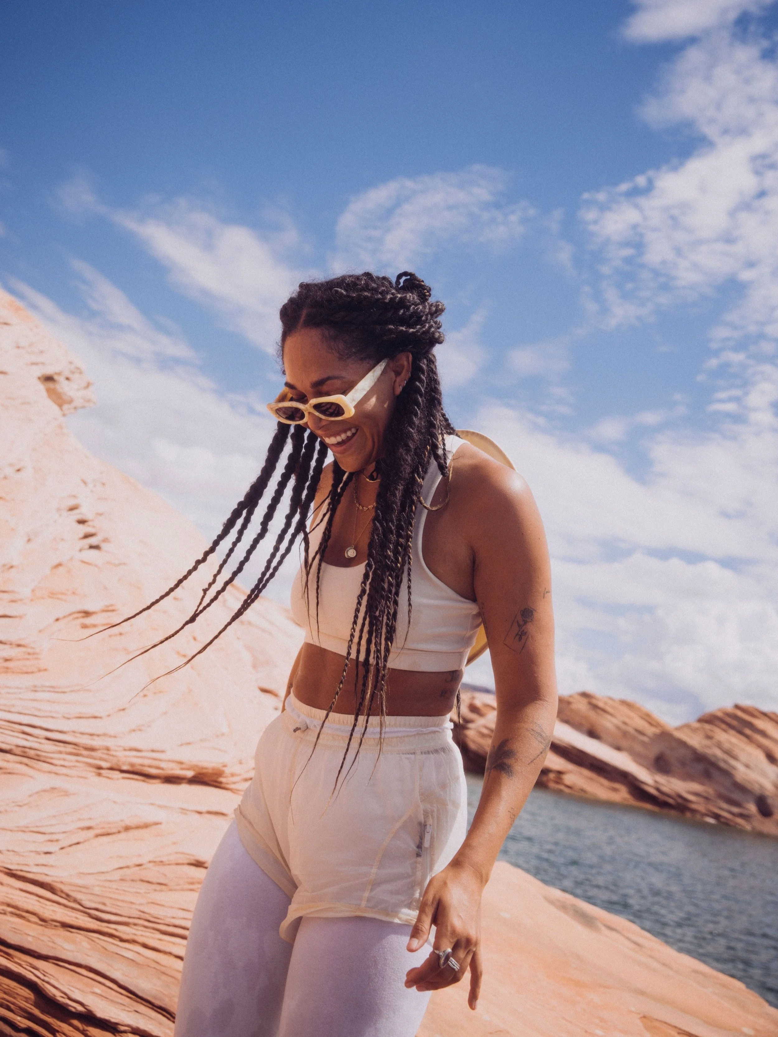 Woman in white activewear with braided hair and sunglasses standing on rock near water.