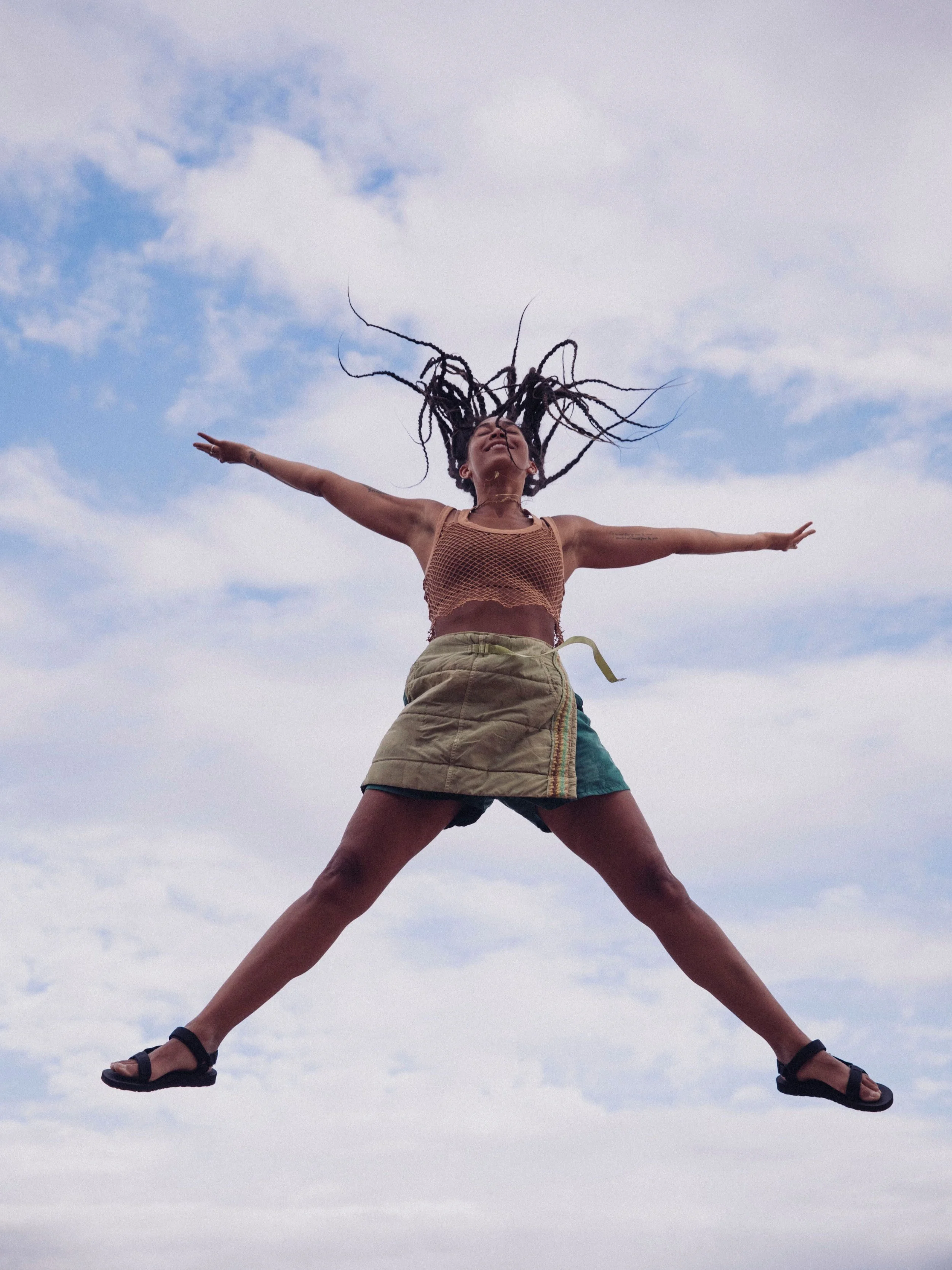 Person with braided hair jumping in the air against a blue sky, wearing a sleeveless top, shorts, and sandals.