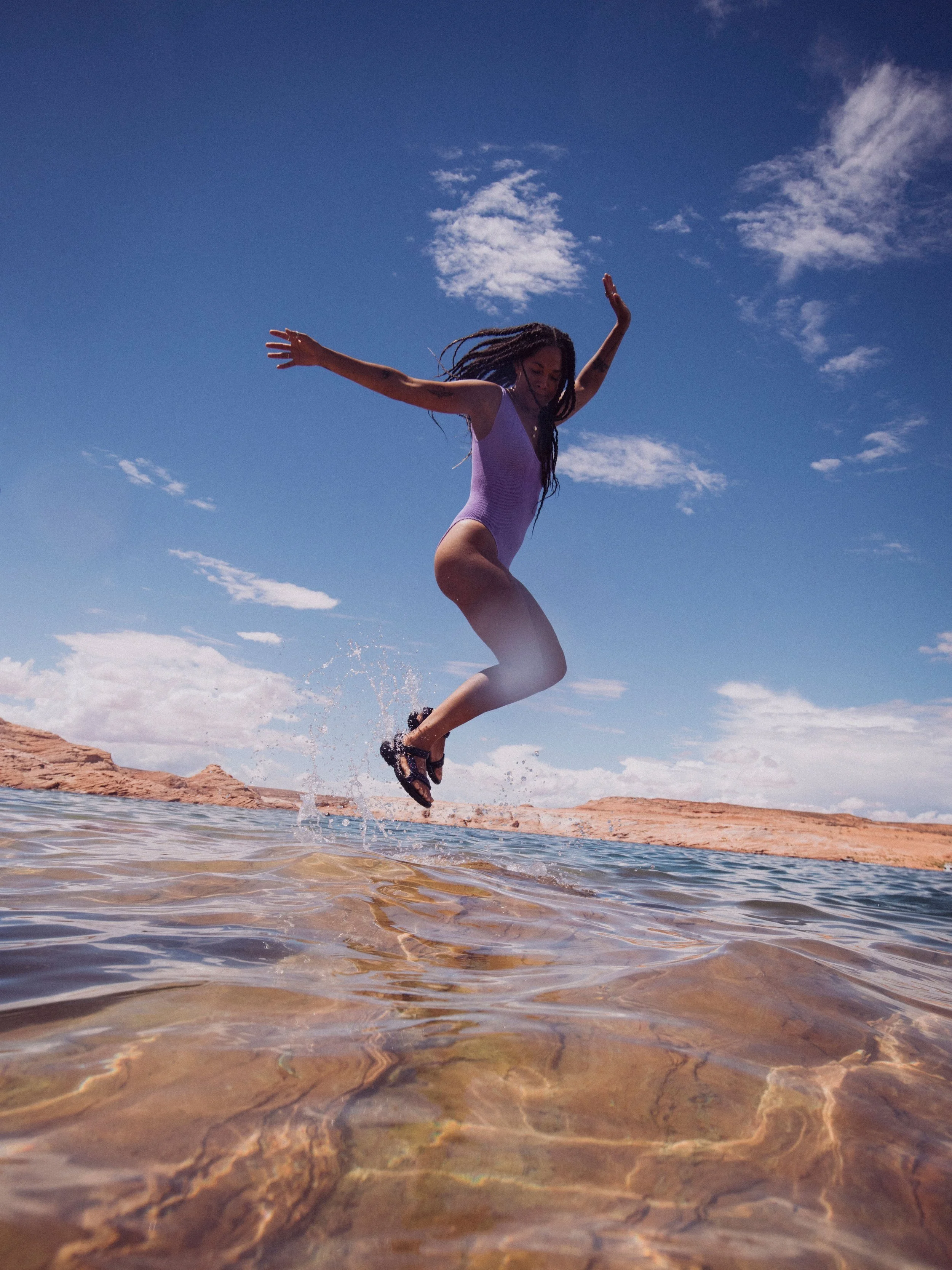 Person in purple swimsuit jumping in water at a beach with blue sky and clouds.