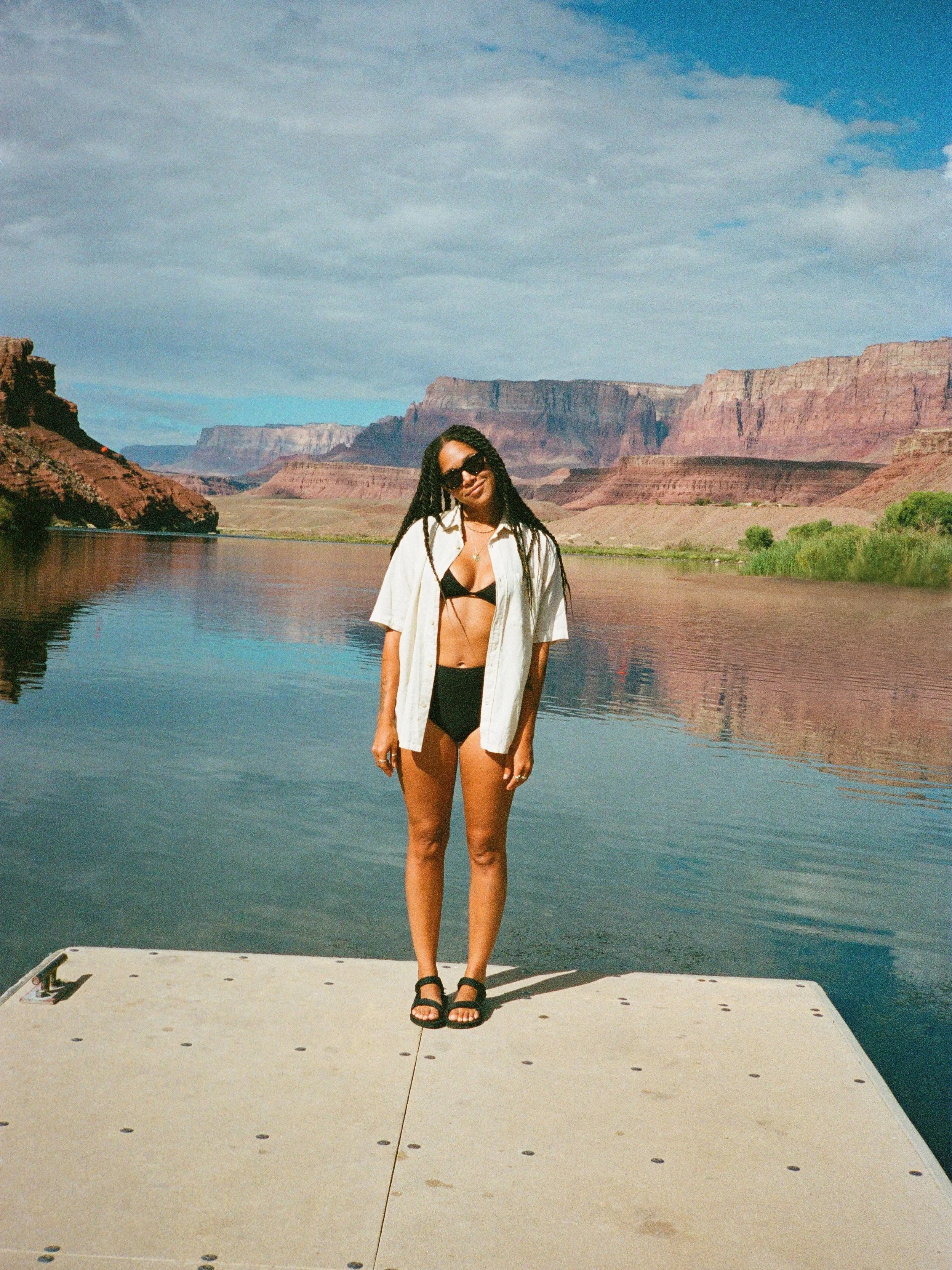 Woman in sunglasses and swimsuit standing on a dock with a scenic background of red rock formations and a river.