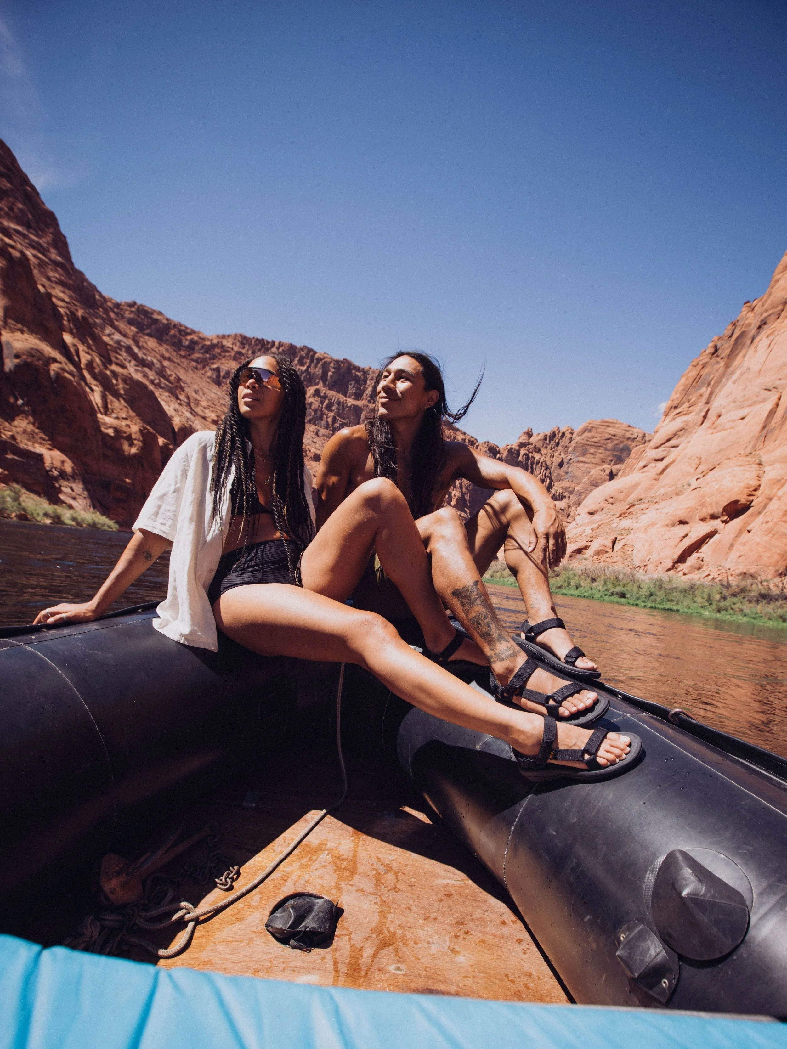 Two people sitting on a black inflatable boat in a canyon, wearing summer clothing and sunglasses, with rocky reddish cliffs in the background under a clear blue sky.