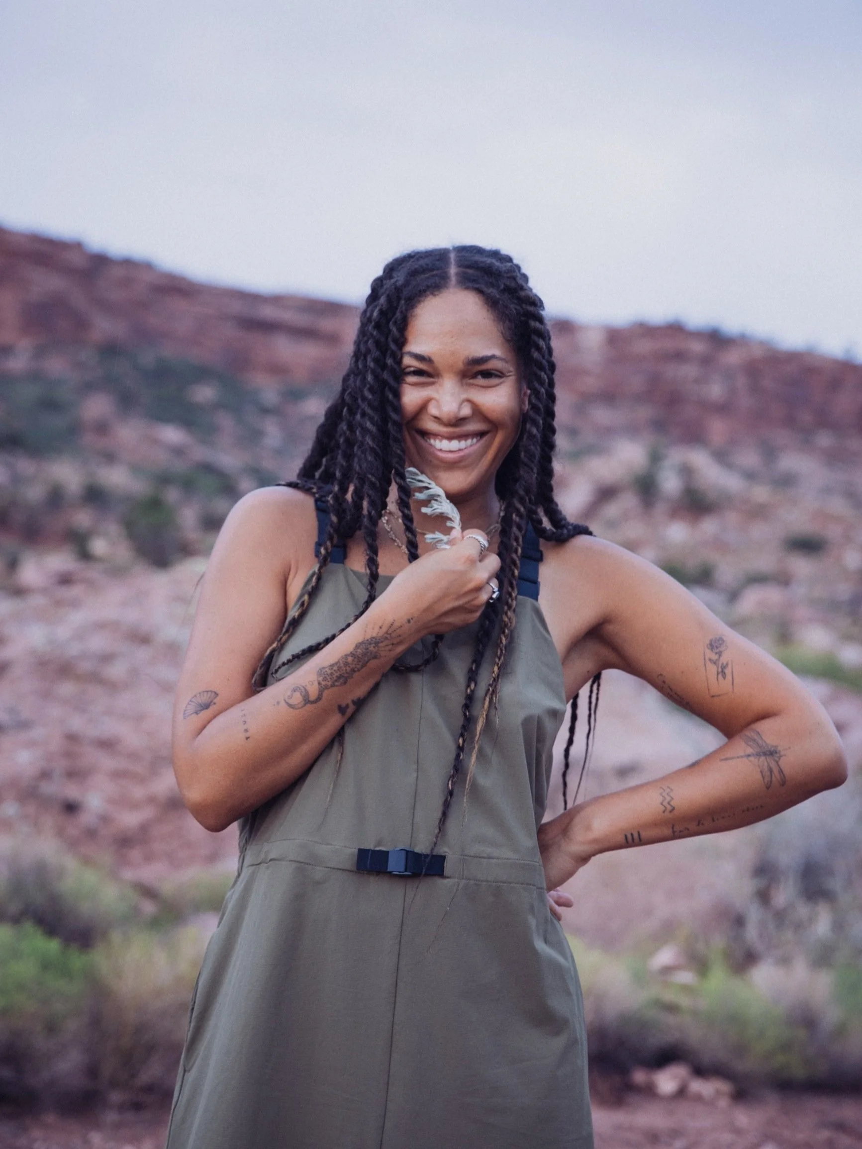 A person with braided hair and tattoos, wearing a green outfit, smiling and holding a feather in a desert landscape.