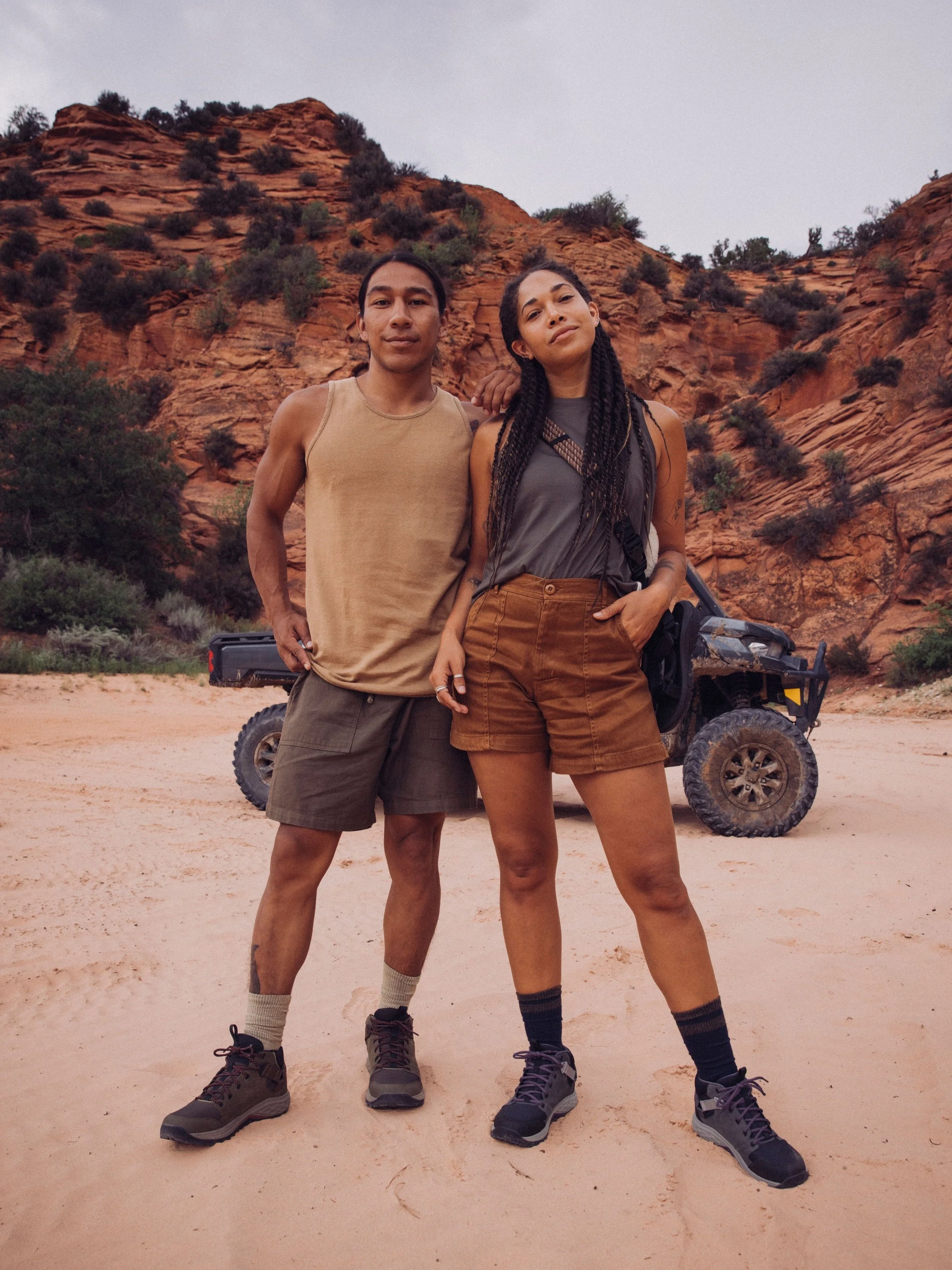 Two people standing in a desert landscape, wearing casual outdoor clothing, with a rocky terrain and an off-road vehicle in the background.