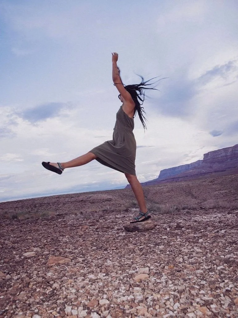 Person in a dress jumping with arms raised on a rocky terrain under a cloudy sky.