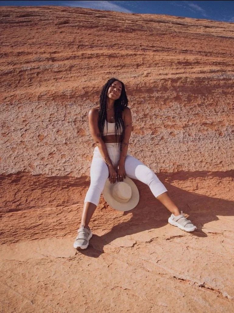 Person sitting on a rocky surface wearing a white outfit, holding a straw hat, with a clear blue sky in the background.