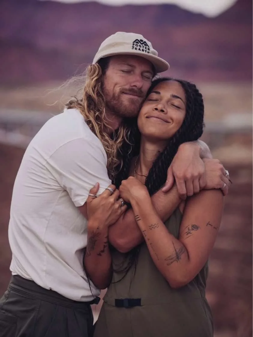 A couple embracing outdoors with tattoos visible on their arms, wearing casual clothing.