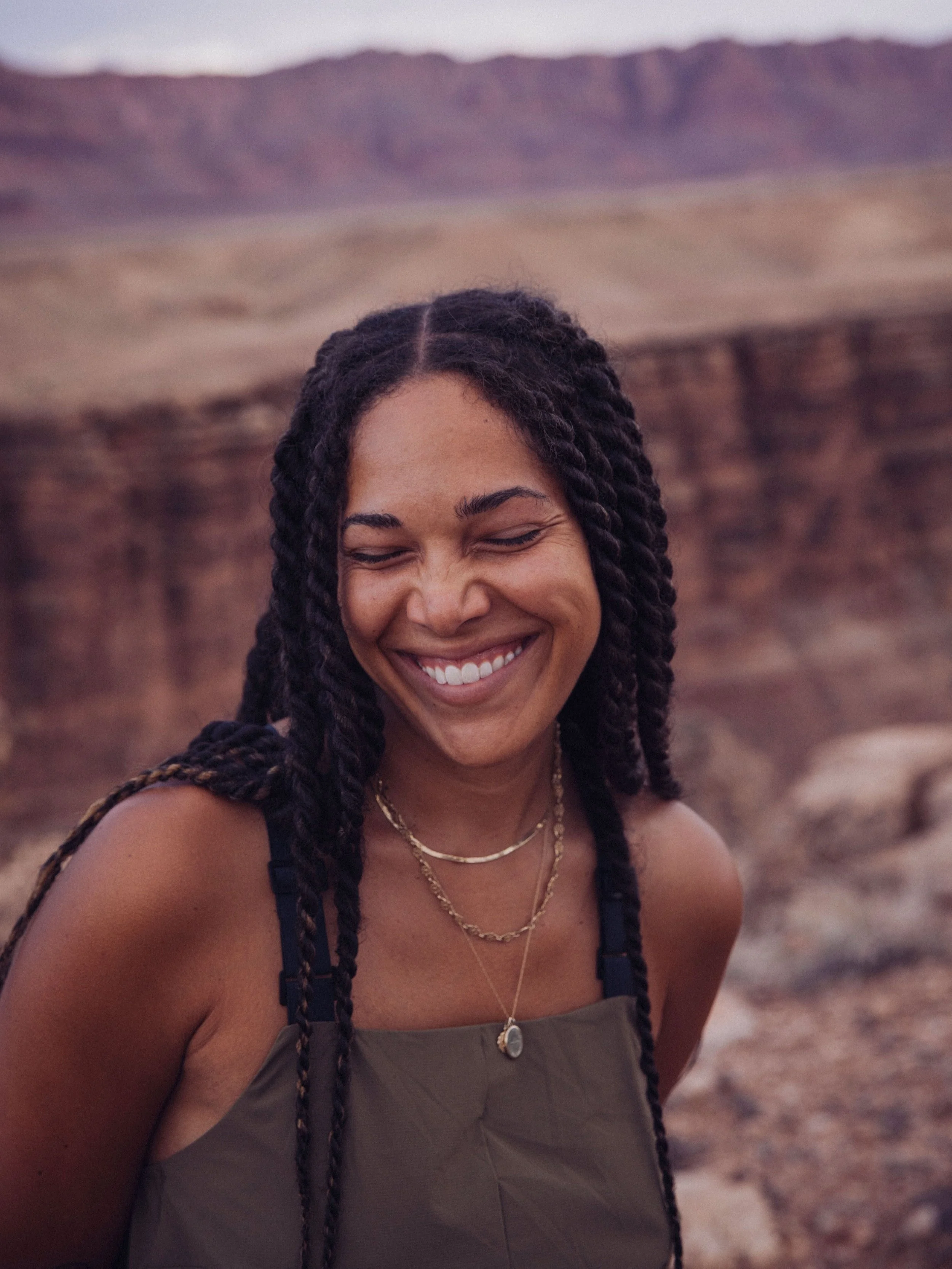 Person with braided hair smiling outdoors in a desert landscape.