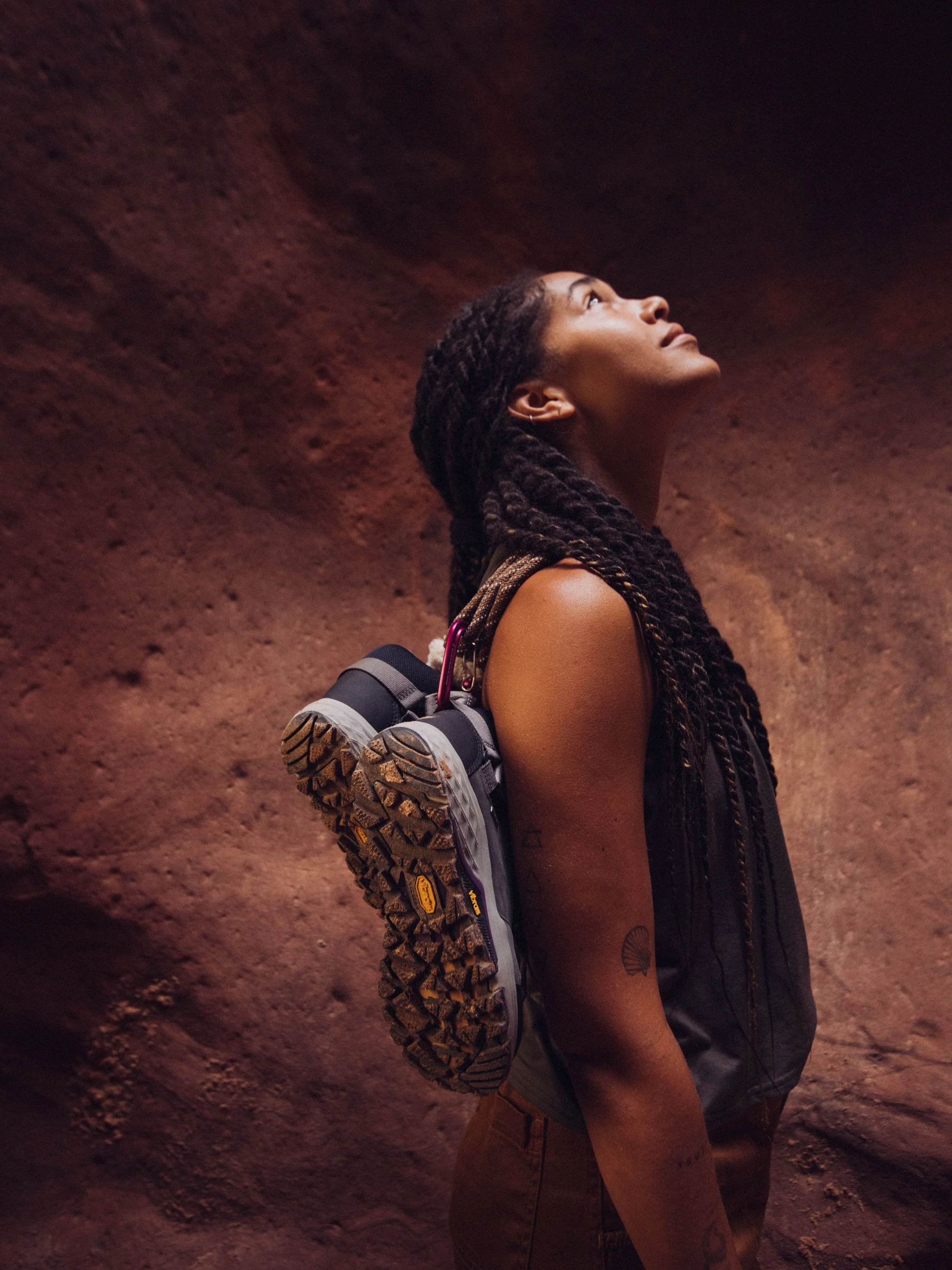 Person with braided hair looking upwards in a rocky environment, with hiking shoes attached to their back.