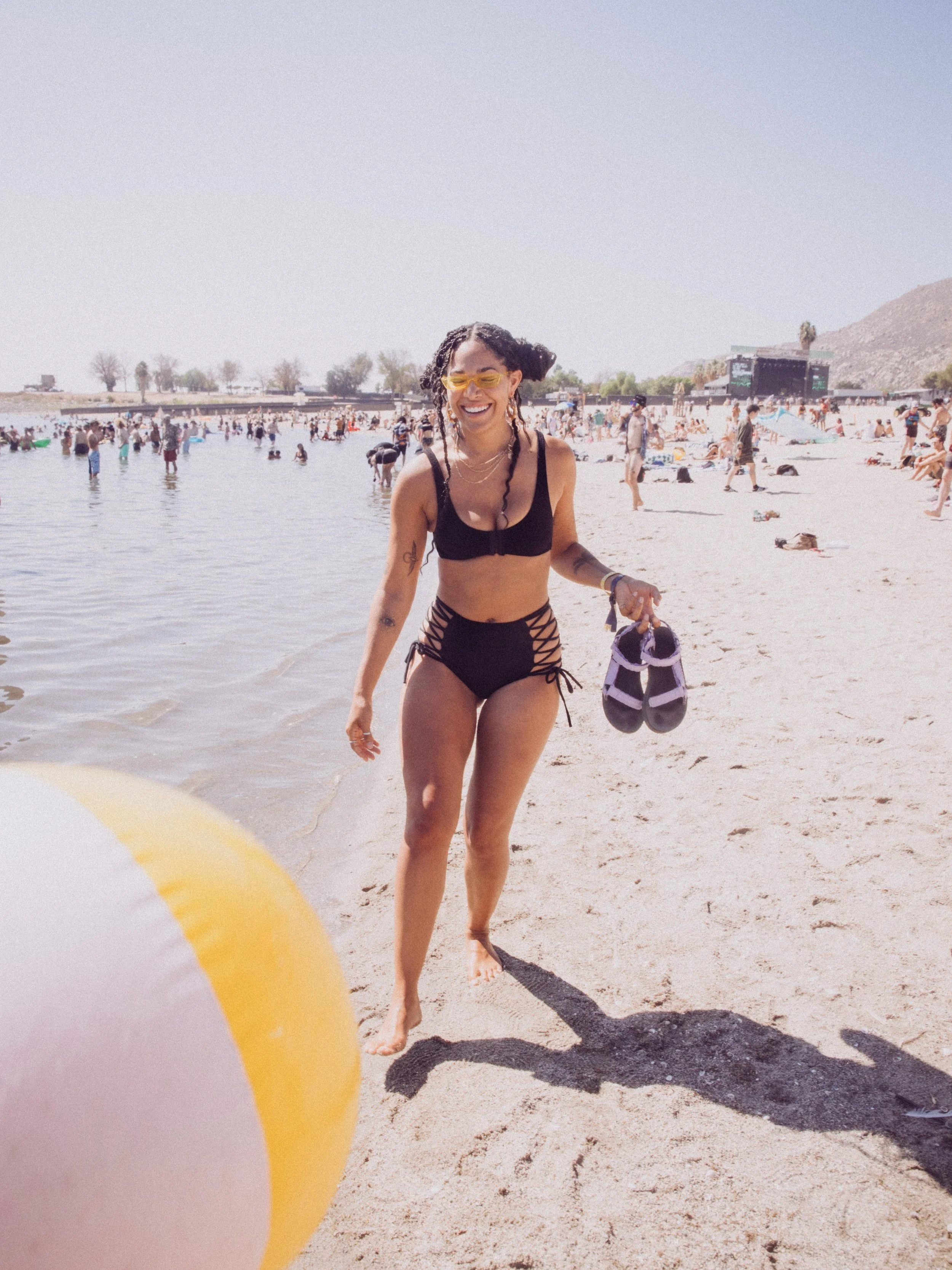 Woman in a black swimsuit on a crowded beach with a yellow beach ball and holding sandals.