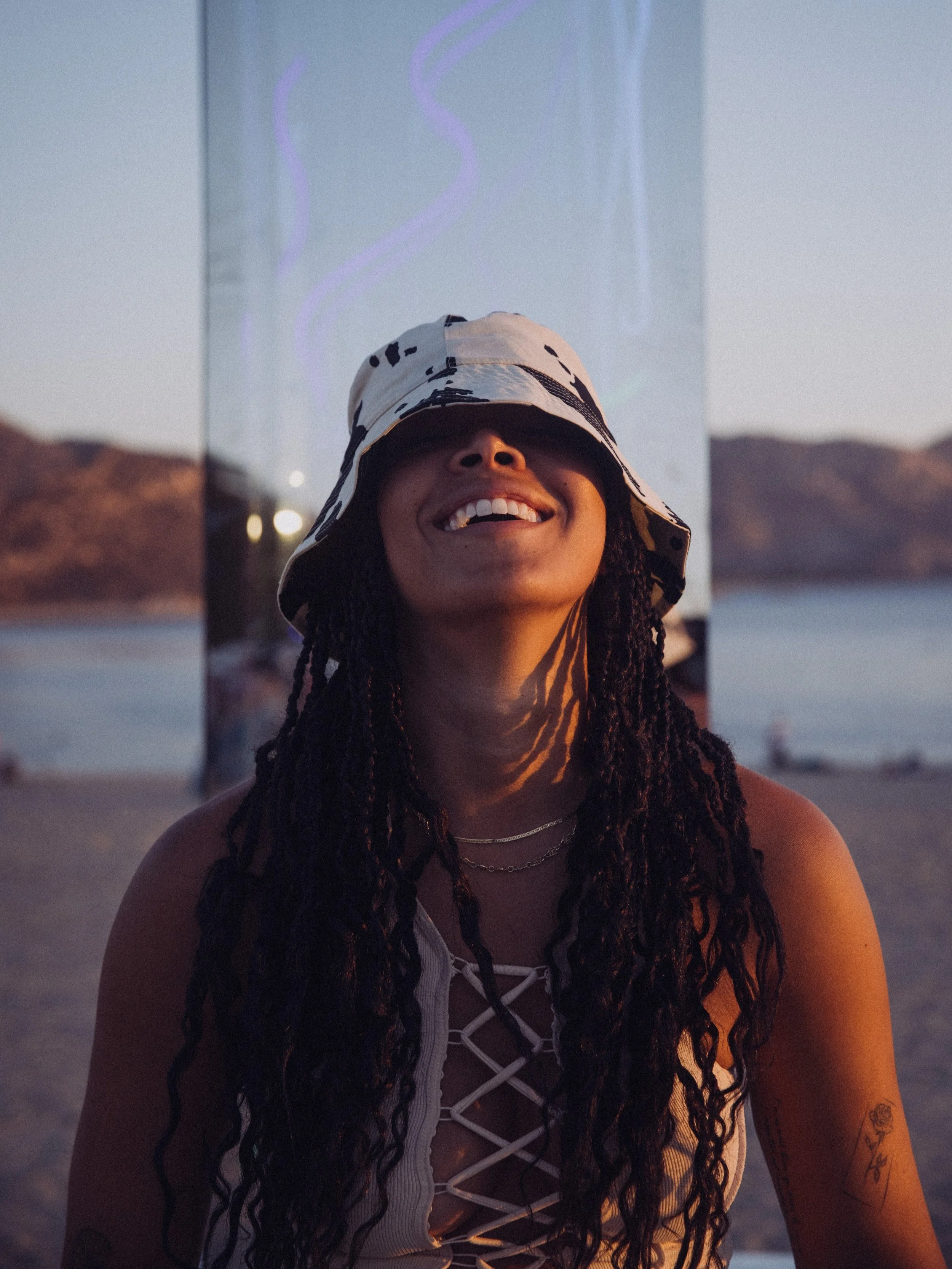 Person wearing a bucket hat and white lace-up top, smiling in front of a beach with mountains in the background.