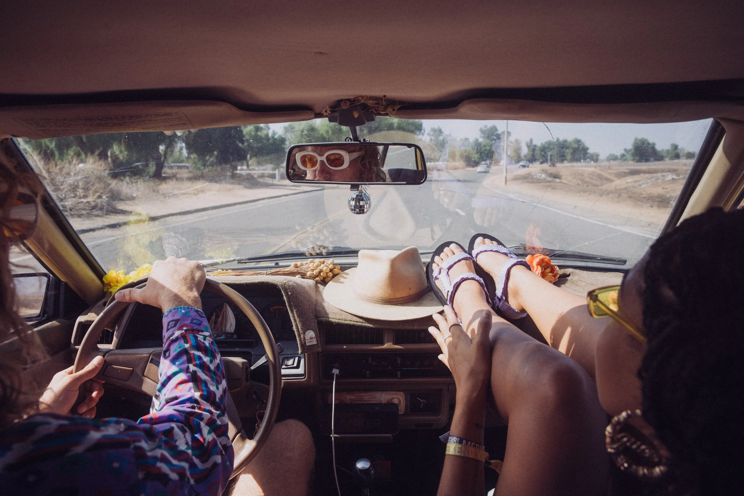 Inside a car, a person is driving with a patterned shirt and sunglasses reflected in the rearview mirror. Another passenger has feet up on the dashboard, wearing sandals, with a hat placed on the dashboard. The road and trees are visible through the 