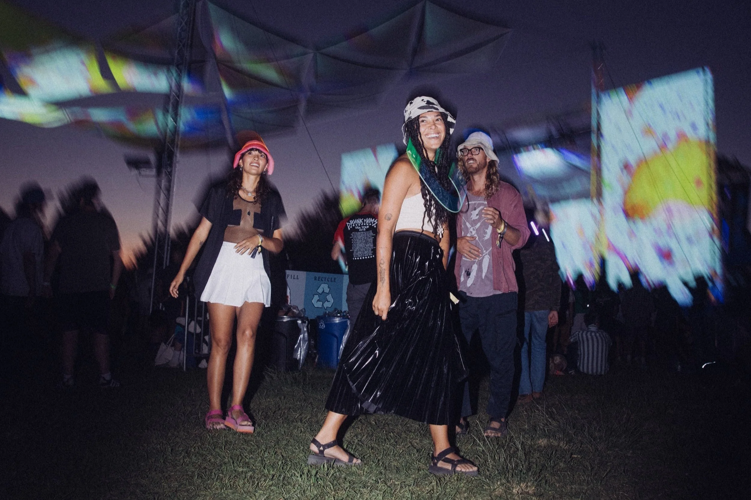 People dancing at an outdoor music festival at night, with colorful projections in the background and wearing casual, festival-style clothing.