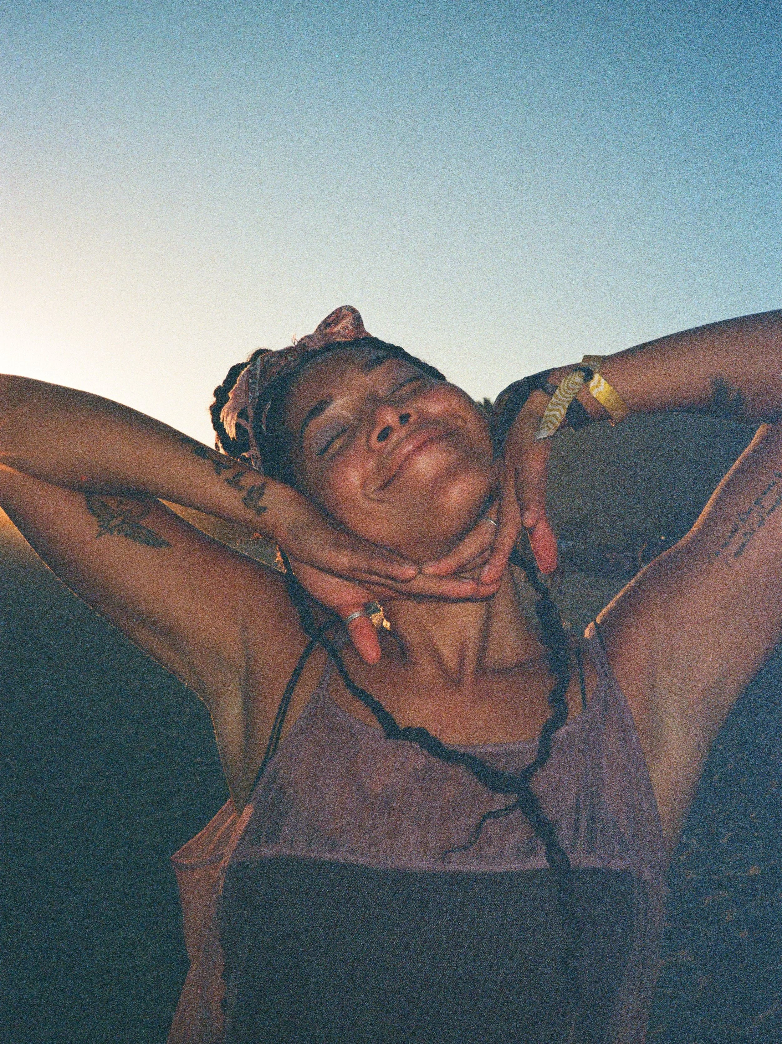 Person smiling with hands on cheeks, wearing a headscarf and sleeveless top, against a sky background.