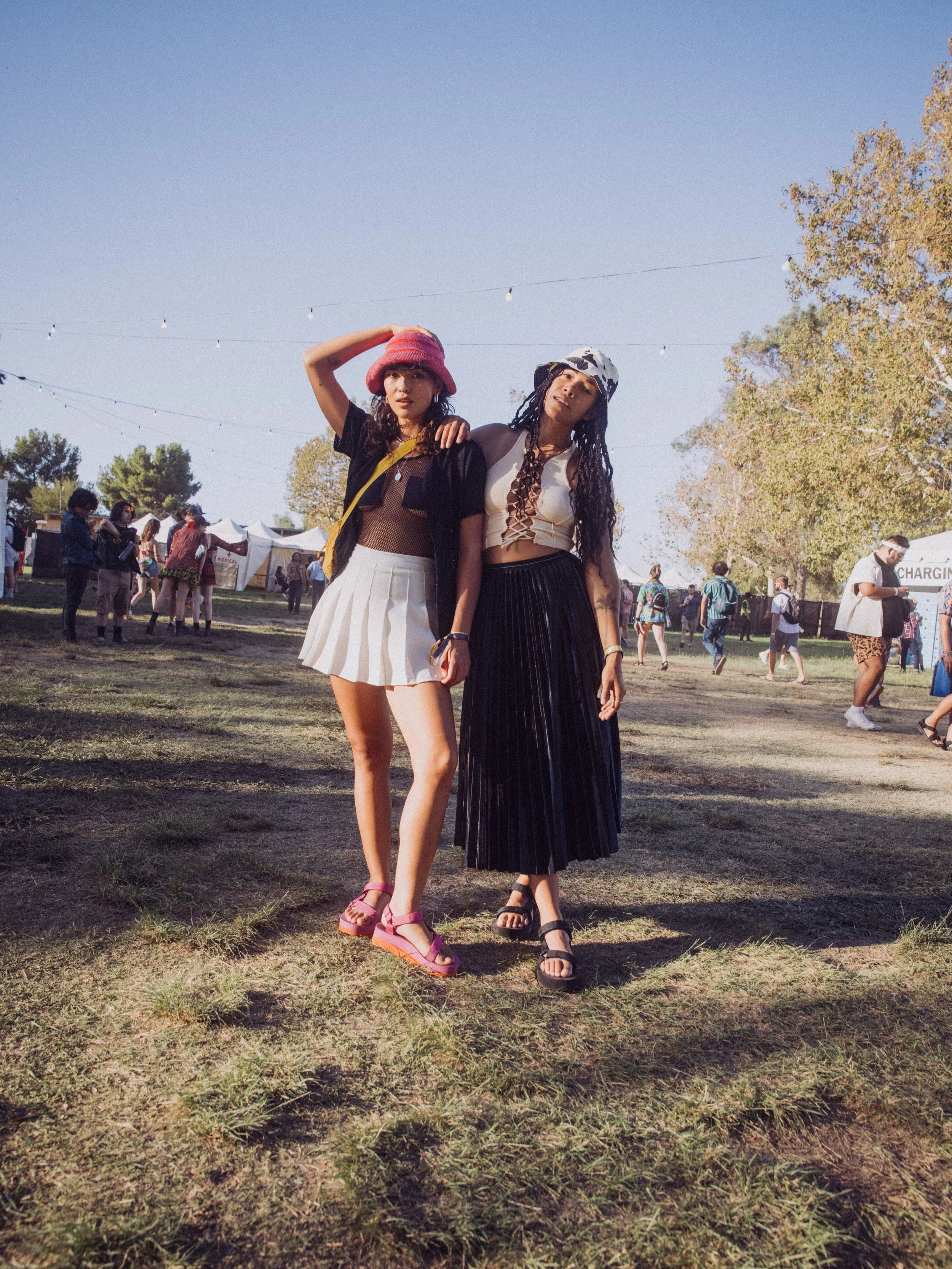 Two people standing on grassy field at outdoor event, wearing casual summer outfits with hats.
