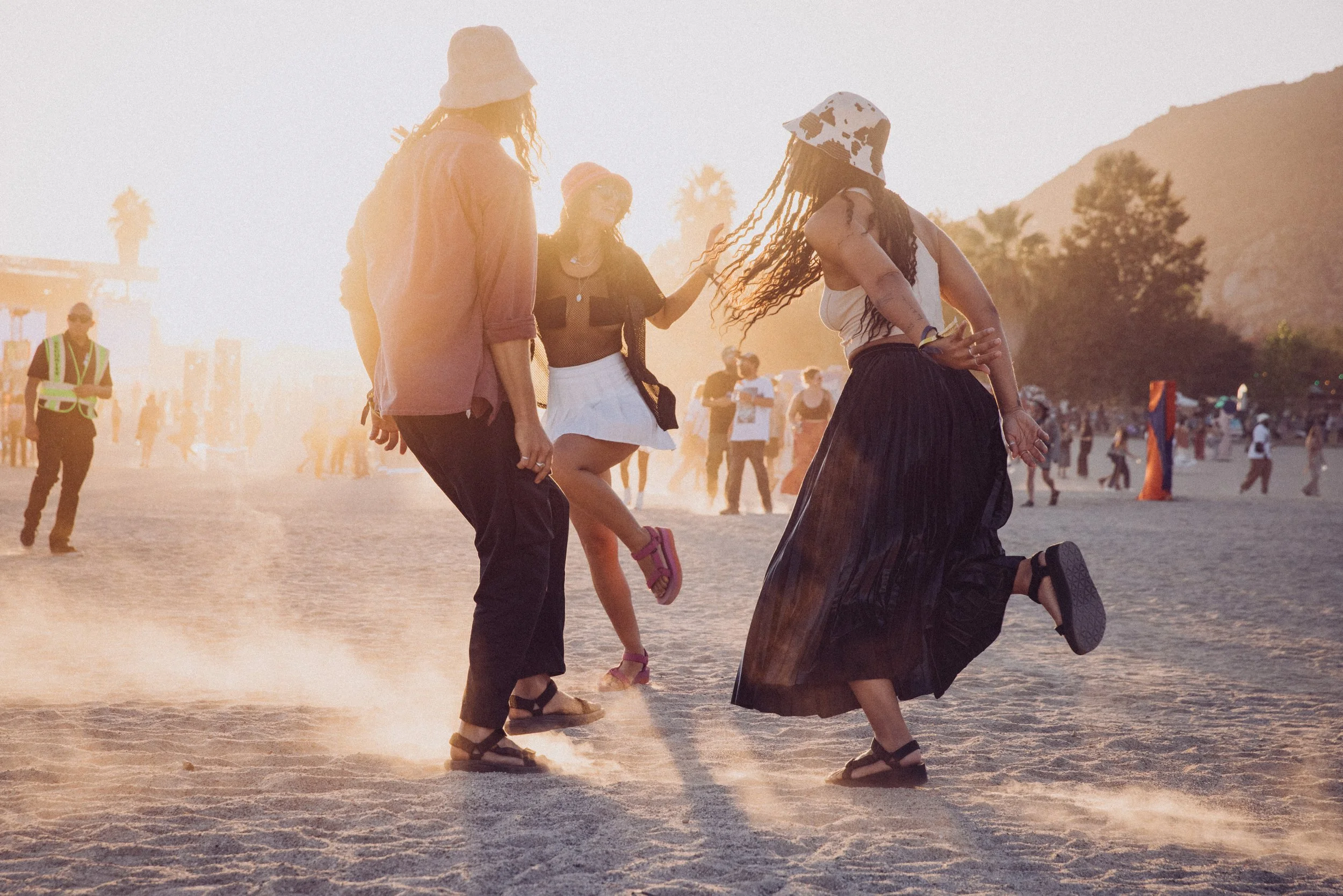 People dancing at a music festival during sunset on a sandy area, wearing casual and festival attire with hats and skirts.