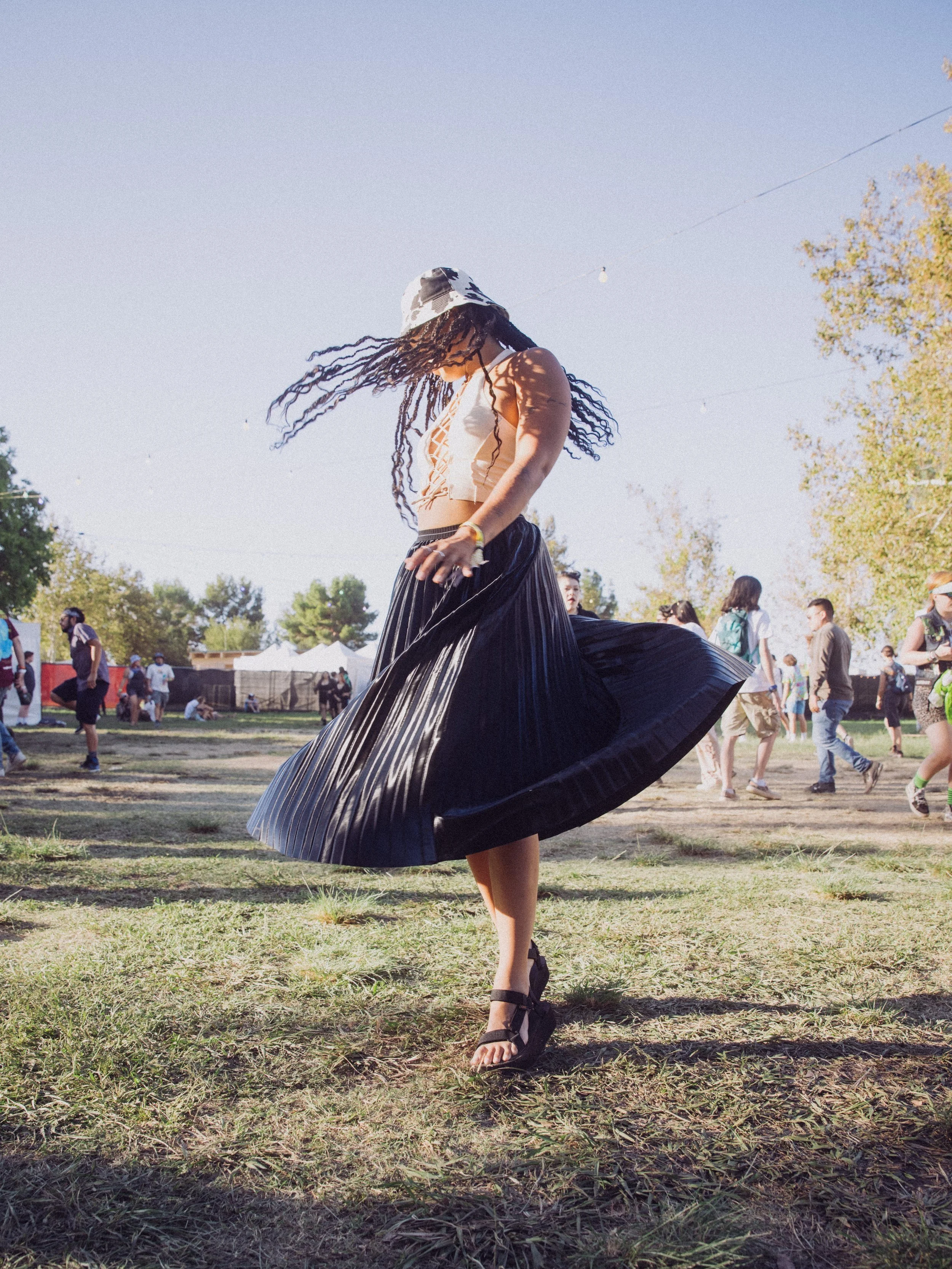 A woman wearing a white crop top, black pleated skirt, and sandals, spinning around on a grassy field at an outdoor event. She has long braids and a bucket hat, with people and trees in the background under a clear blue sky.