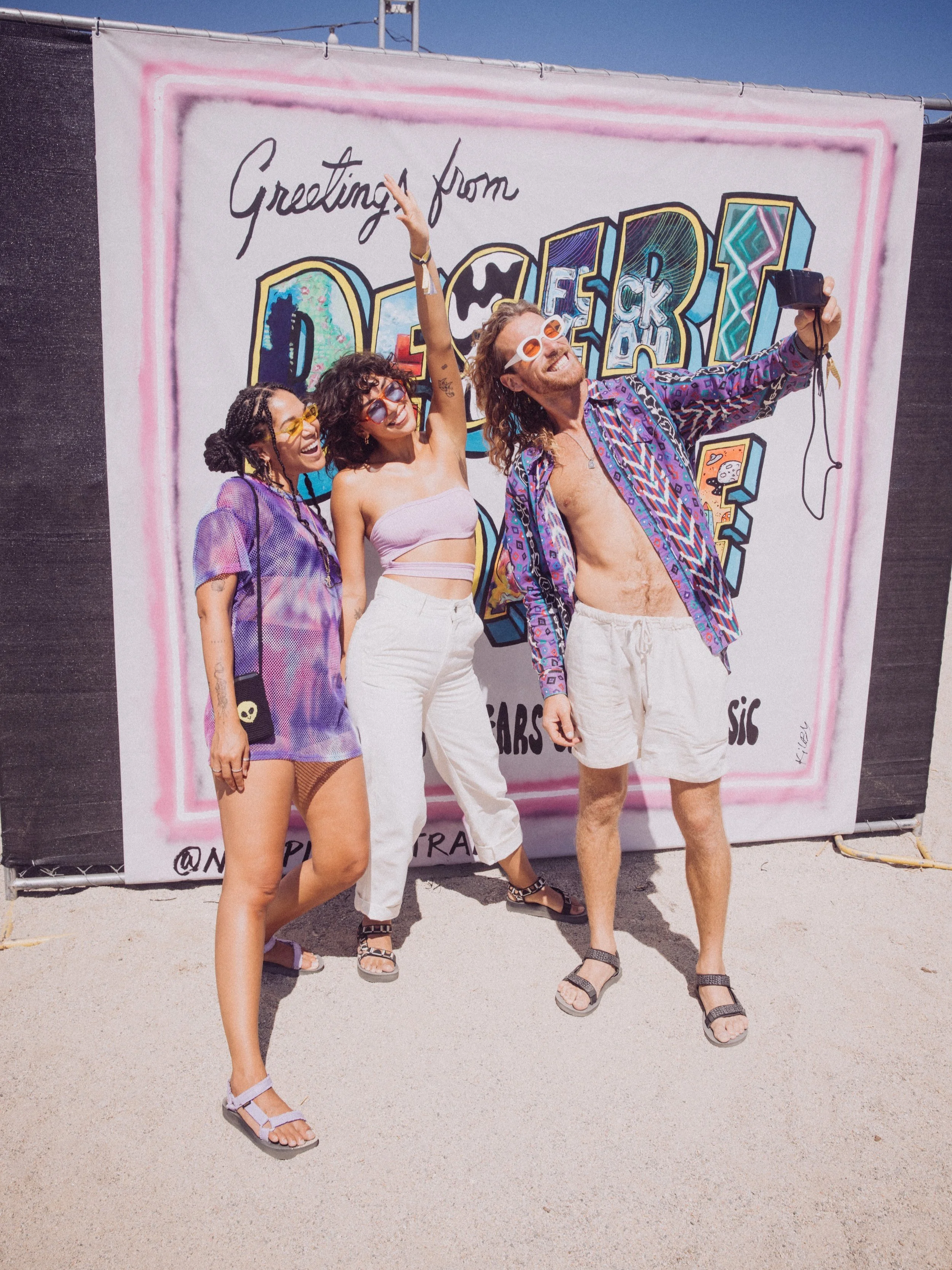 Three people taking a selfie in front of a colorful festival backdrop with "Greetings from Desert" written on it. They are wearing casual summer clothing, sunglasses, and look joyful.