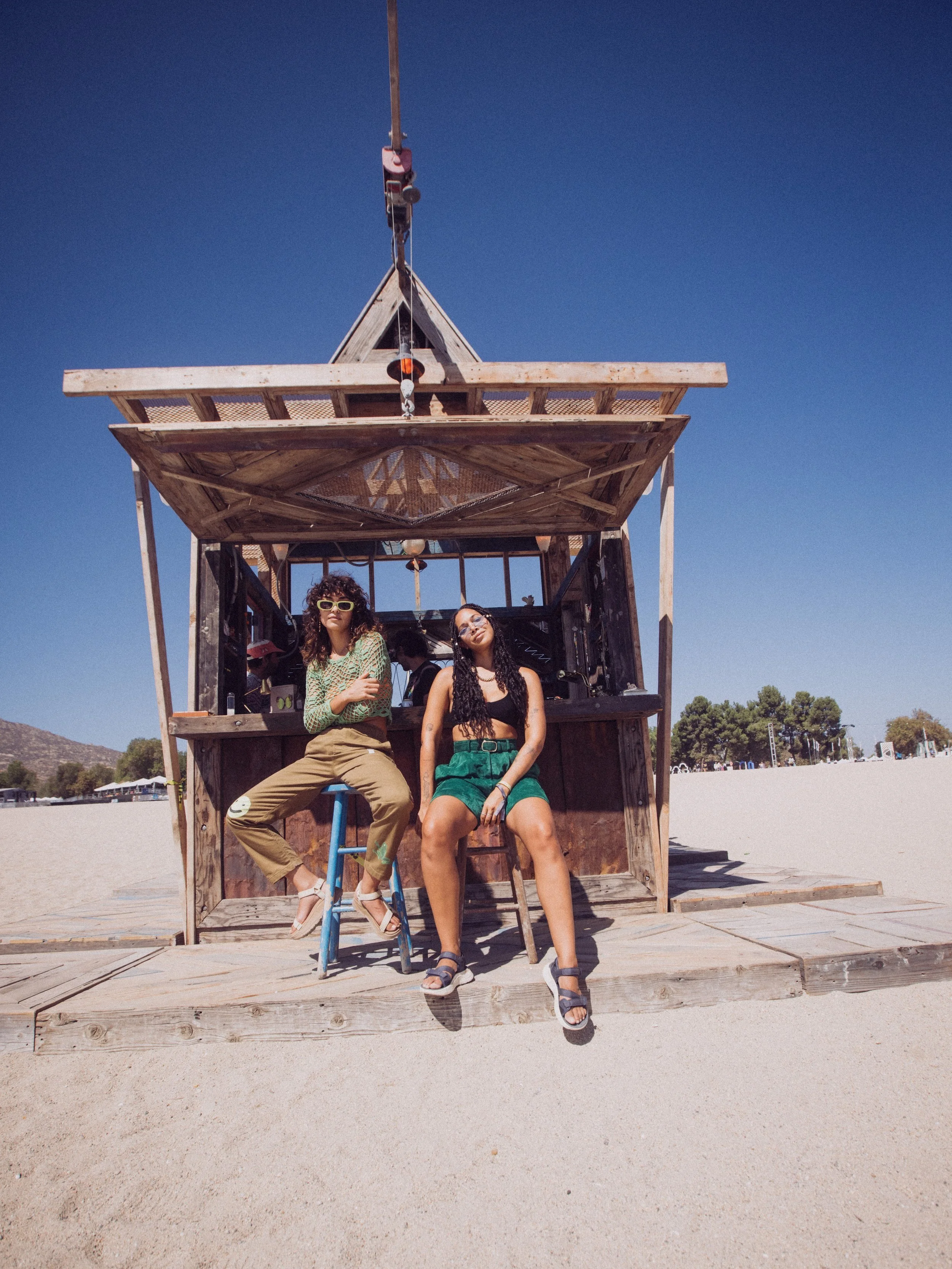 Two people sitting in front of a small wooden structure on a sandy beach, under a clear blue sky.