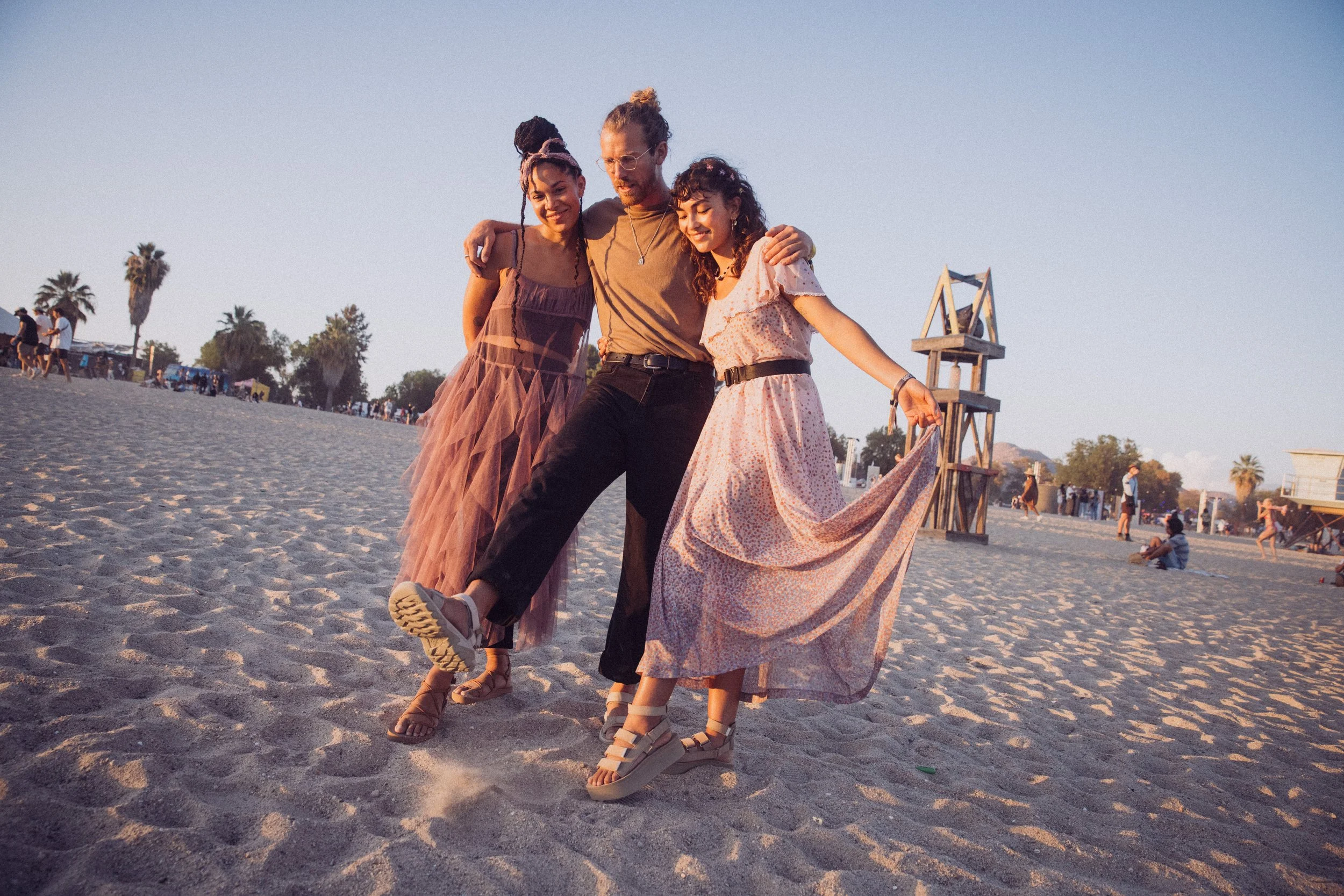 Three people posing together on a sandy beach with palm trees, wearing summery clothing and sandals, during sunset. A wooden structure and other people in the background.
