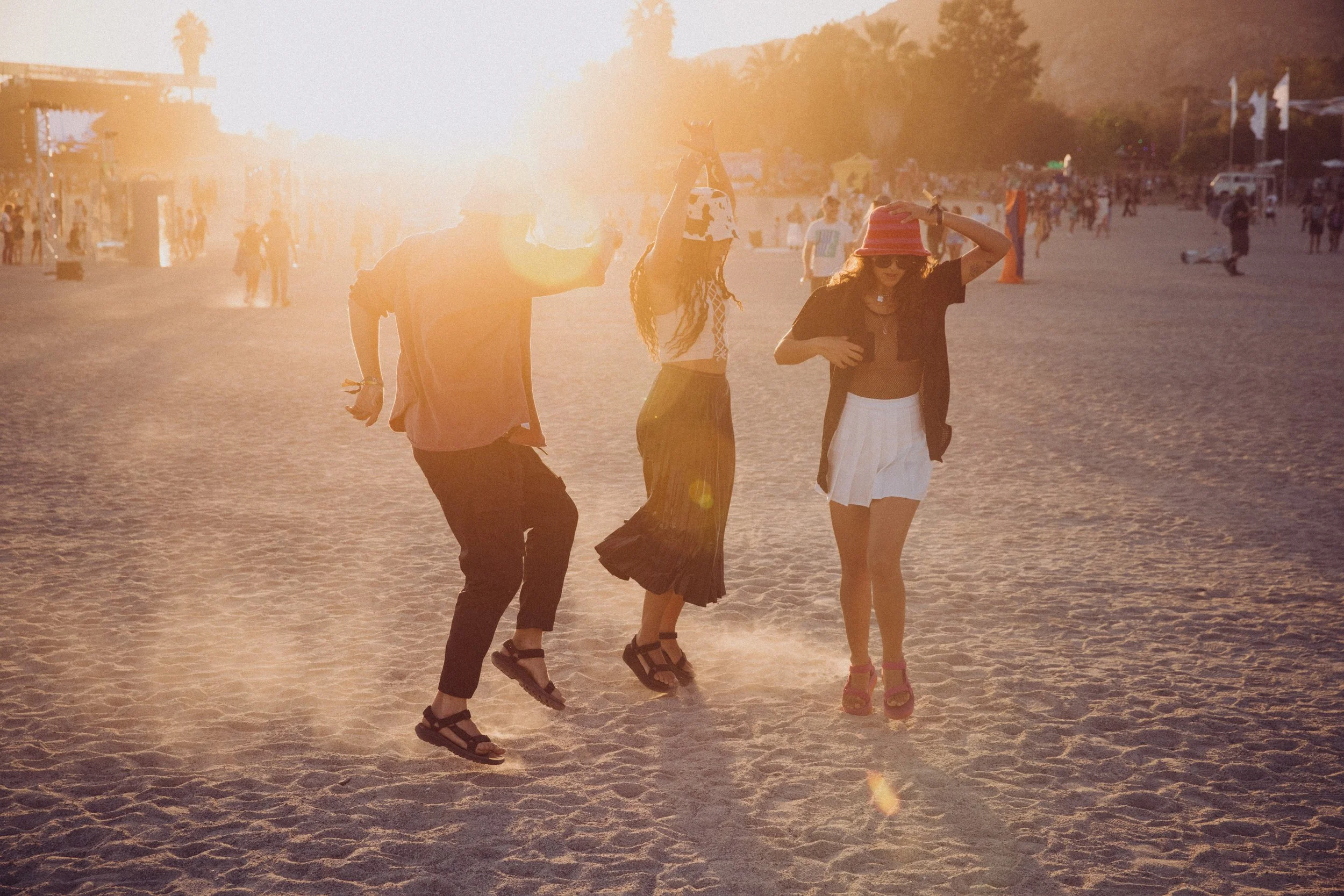 Three people dancing on a sandy beach at sunset with a festival-like atmosphere.