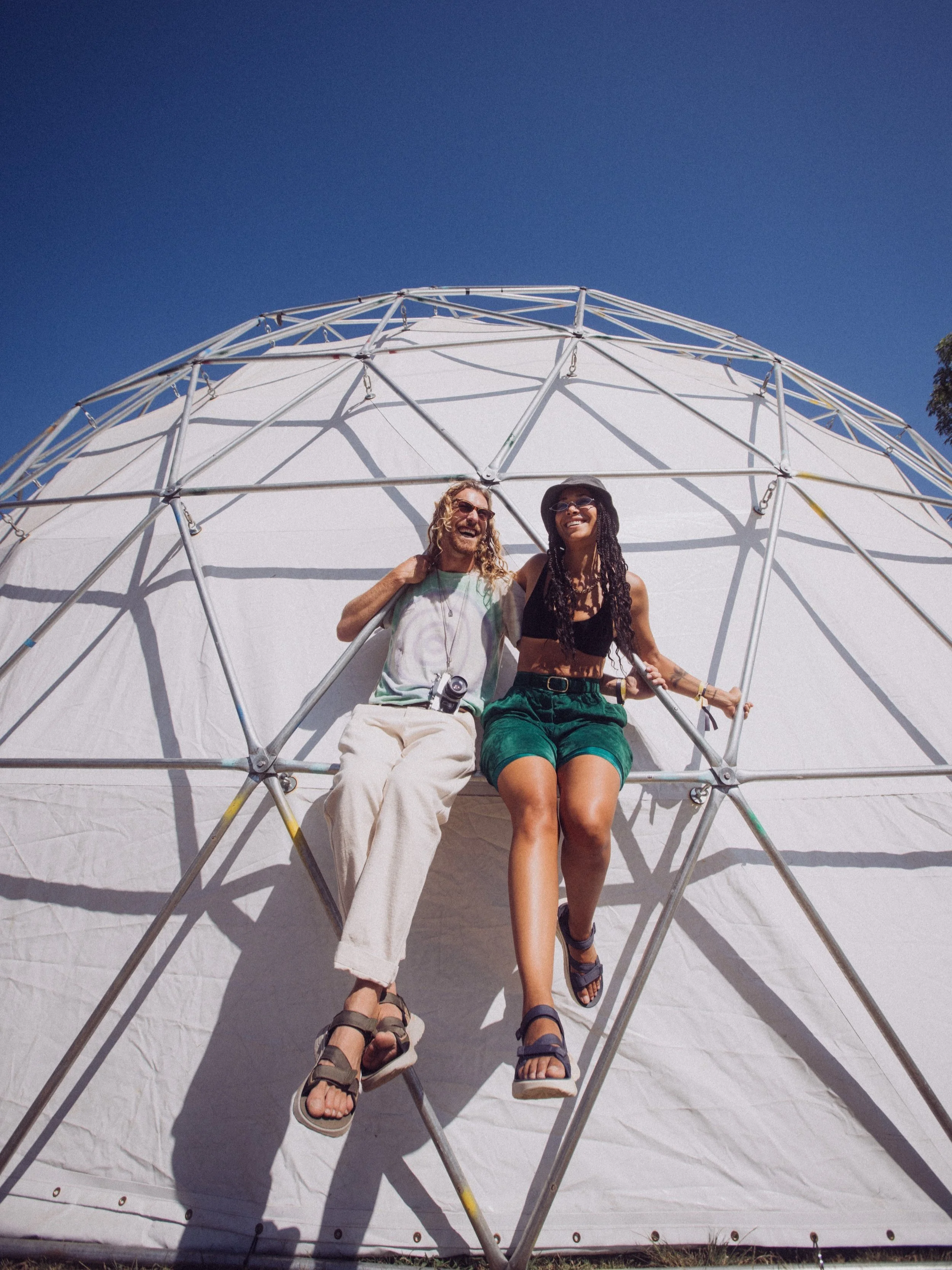 Two people sitting on the frame of a geodesic dome against a clear blue sky. One is wearing light-colored pants and sandals, the other in green shorts and dark sandals.
