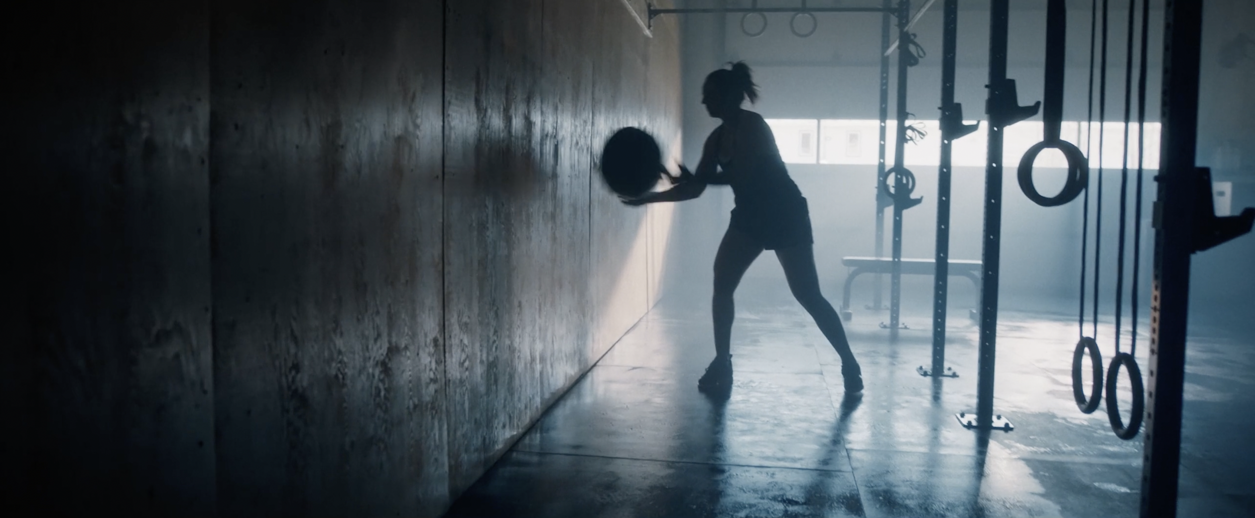 Silhouette of a person working out with a medicine ball against a wall in a gym, with gymnastic rings and exercise equipment in the background.