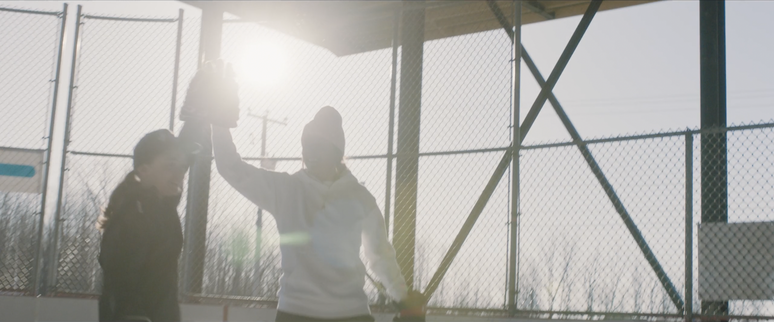 Two people high-fiving on an outdoor ice rink with sunlight shining through a chain-link fence.