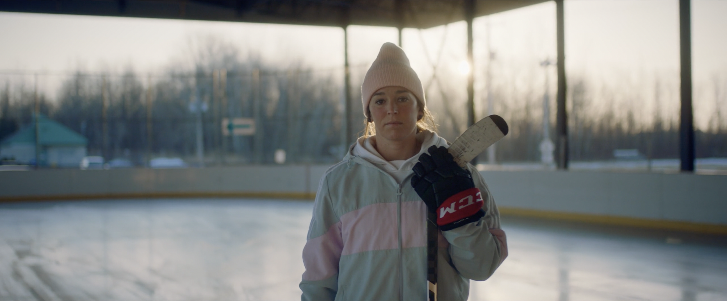 Person in a beanie holding a hockey stick on an outdoor ice rink.