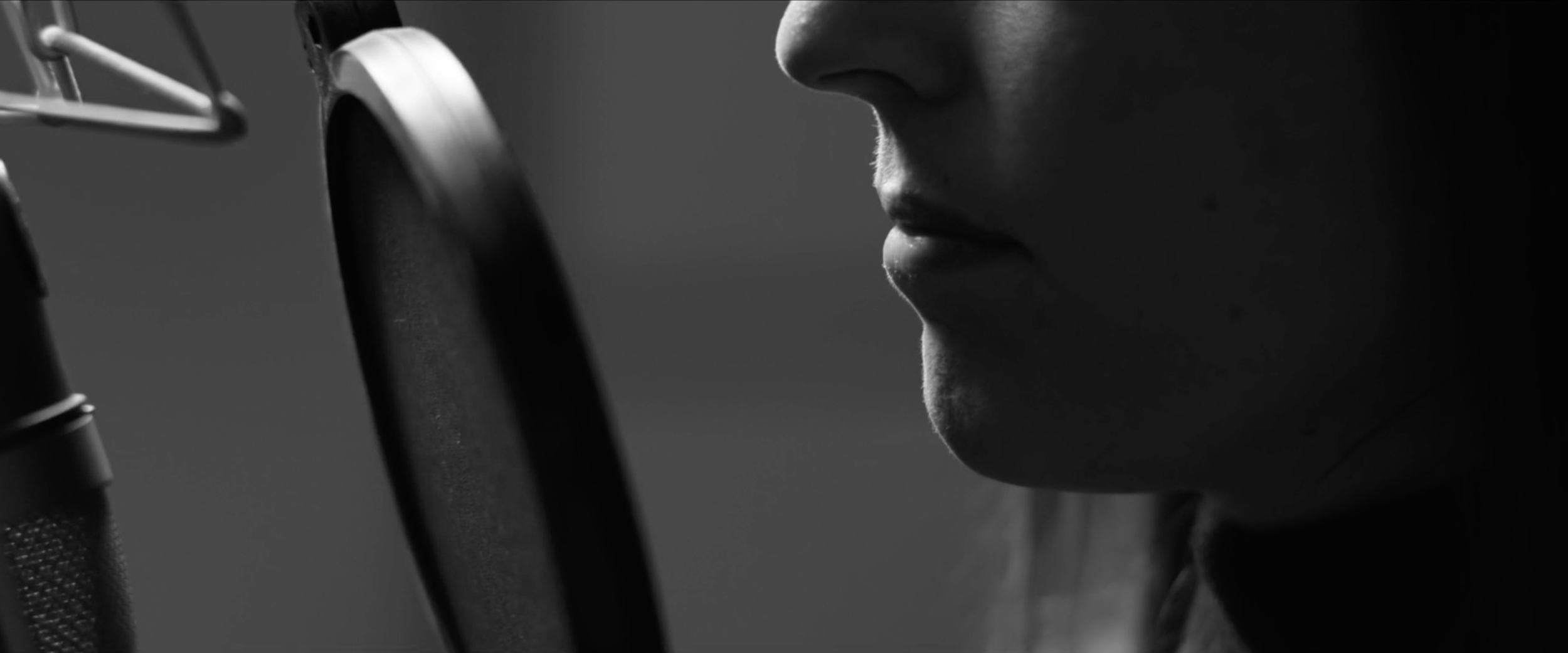 Close-up of a person's mouth speaking into a microphone with a pop filter, in black and white.
