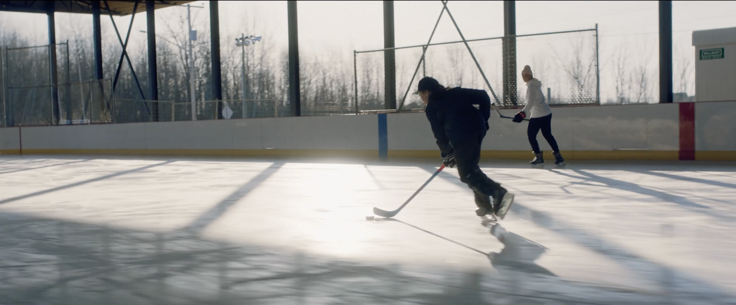 Two people playing ice hockey on an outdoor rink, one in black skating towards the camera with a stick, and the other in white and black following behind.