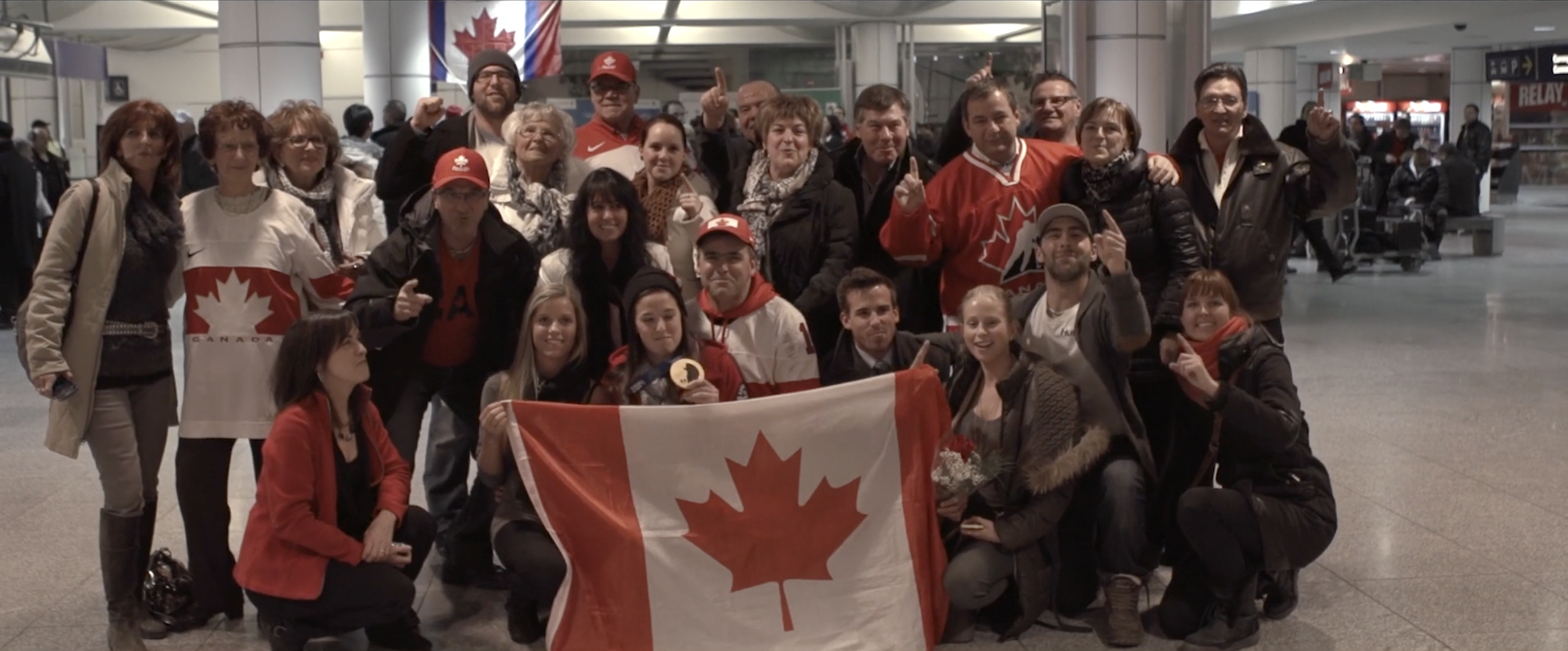 Group of people in an indoor setting holding a Canadian flag, wearing red and white clothing with Canada-themed designs.