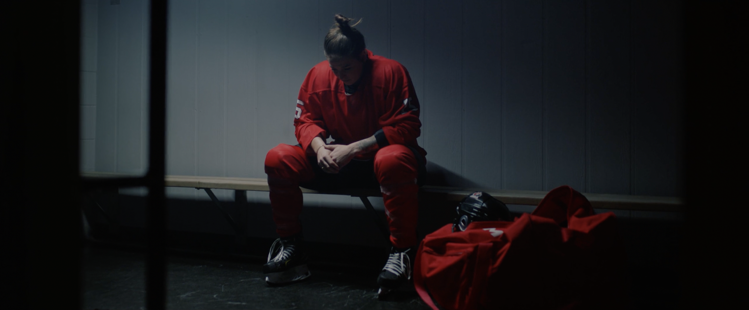 Hockey player in red uniform sitting on a bench, looking down, with a hockey bag and helmet nearby, in a dimly lit locker room.