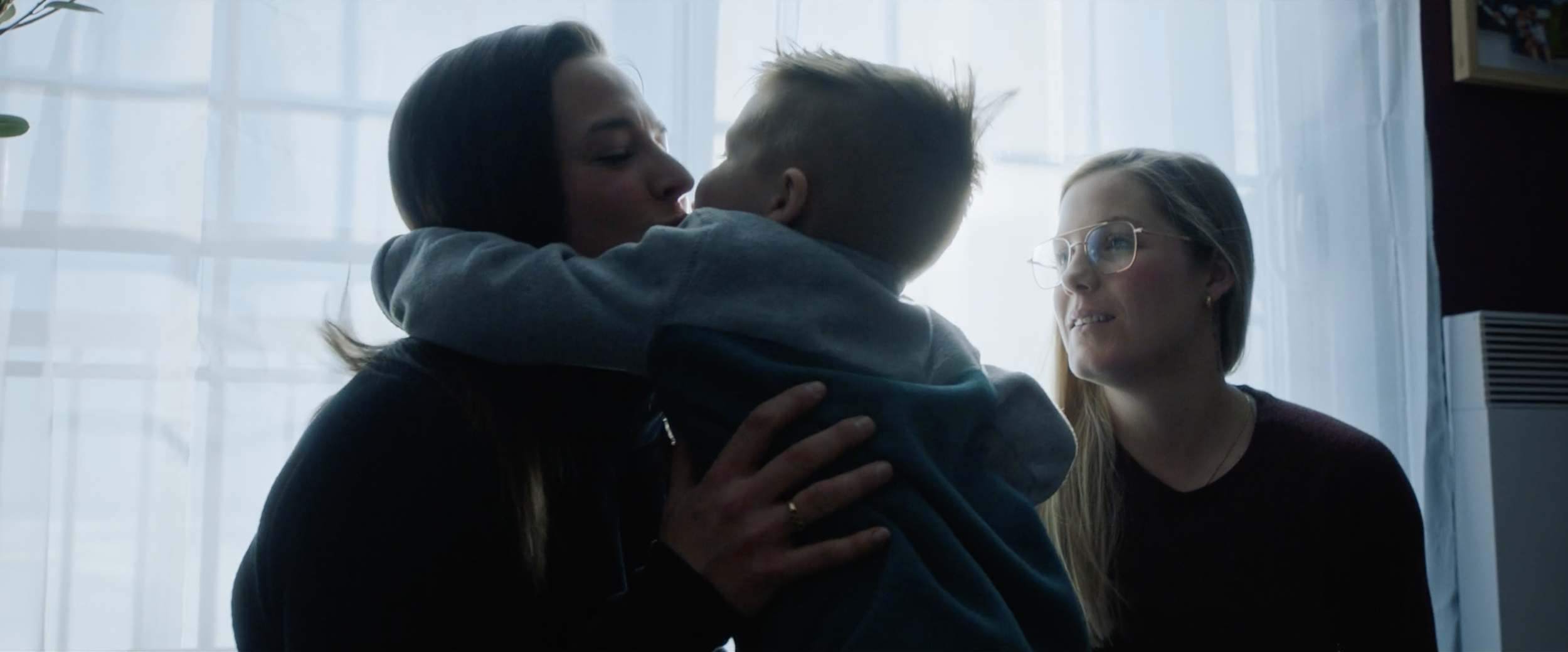 A person hugging and kissing a child while another person with glasses looks on, all in a softly lit room.