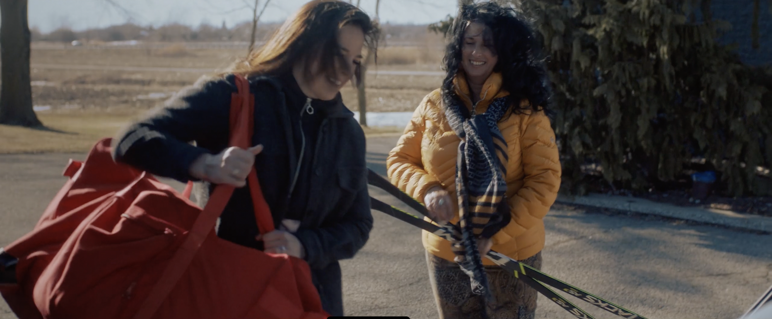 Two women outdoors, one holding a red bag, the other holding skis and wearing a yellow jacket.