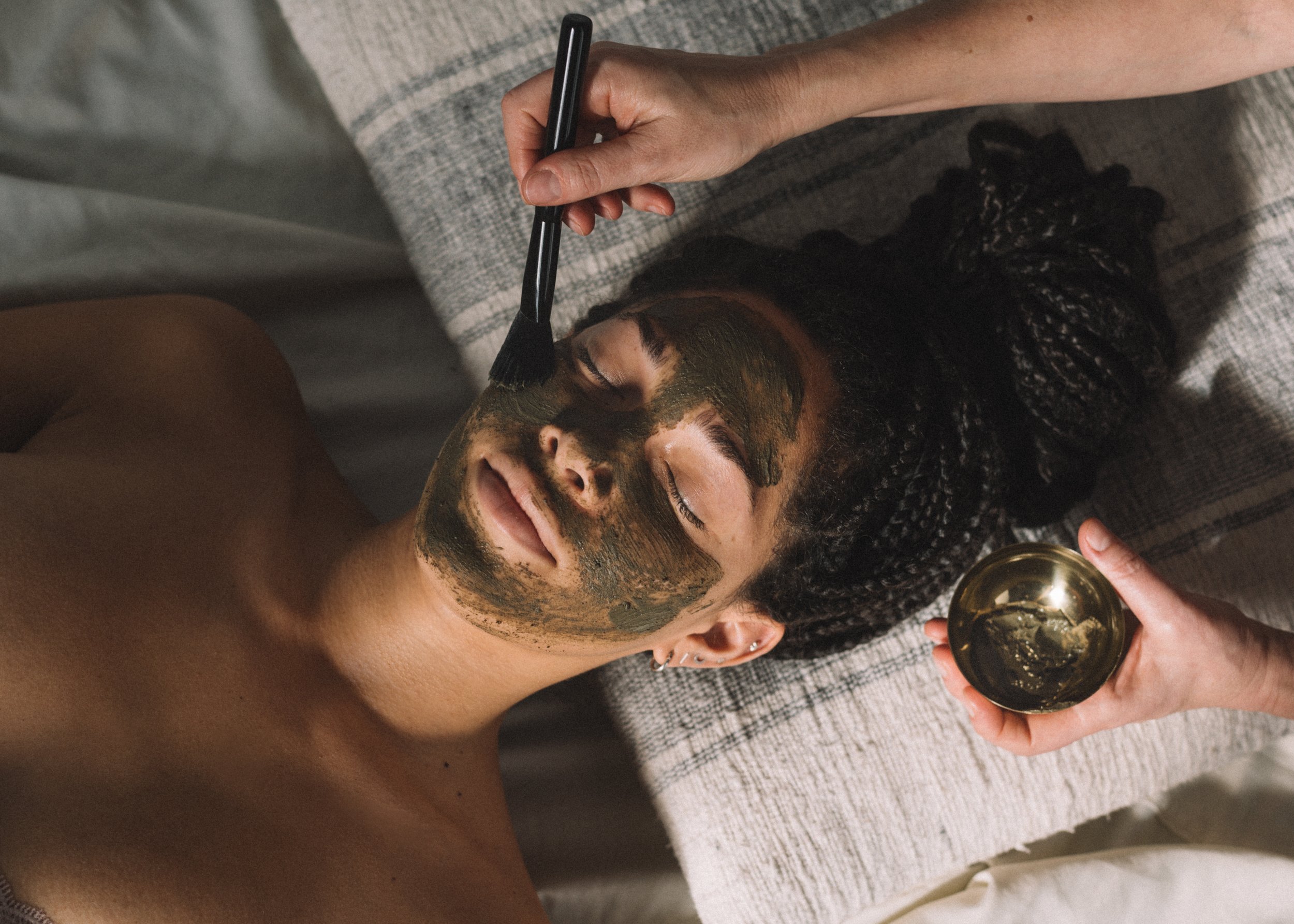 Person receiving a facial treatment with a green mask being applied using a brush, while lying on a towel-covered surface.