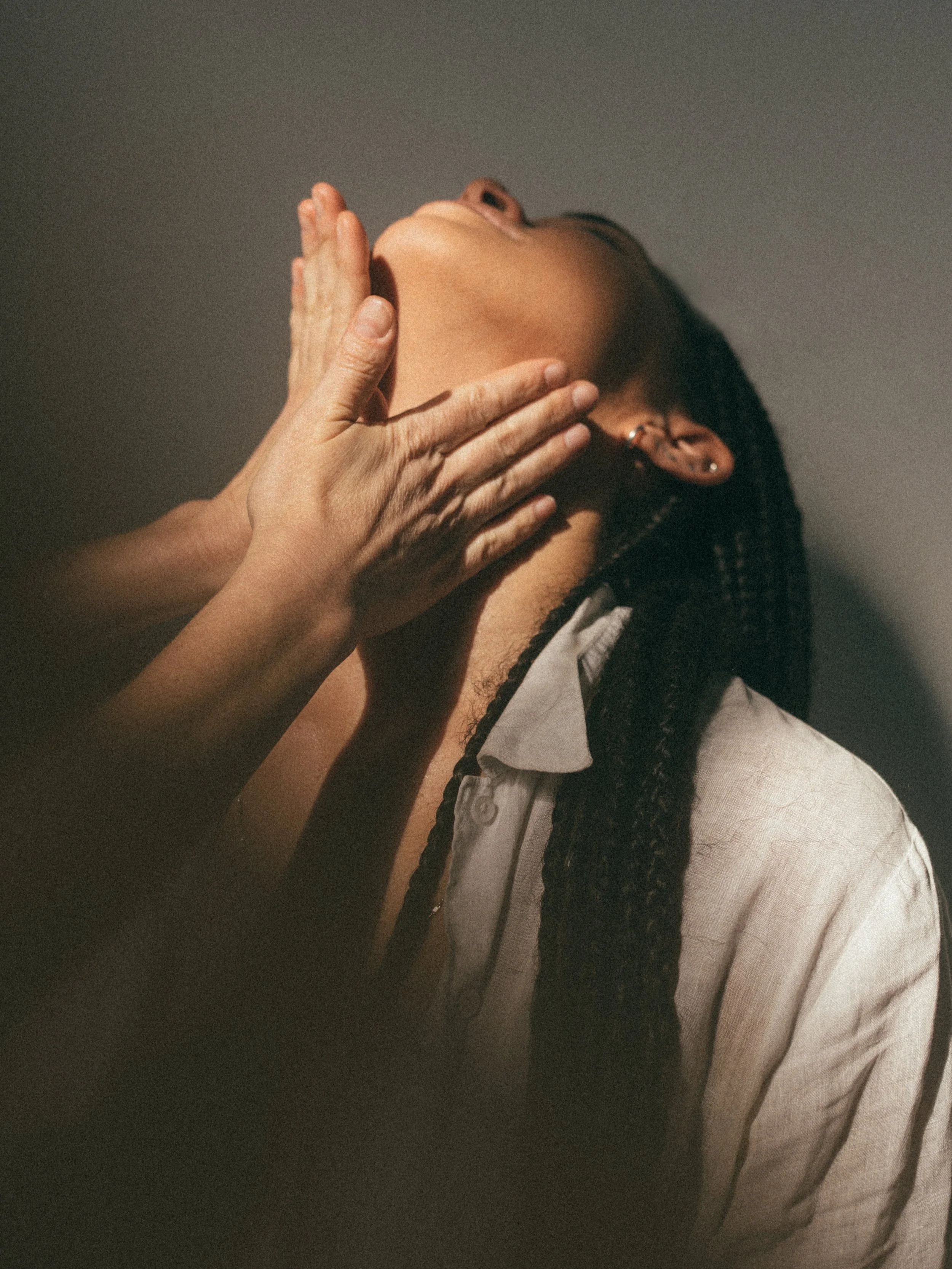 A person with an upturned face and closed eyes receives a facial massage from another person's hands, with long braided hair and a partially visible white shirt.
