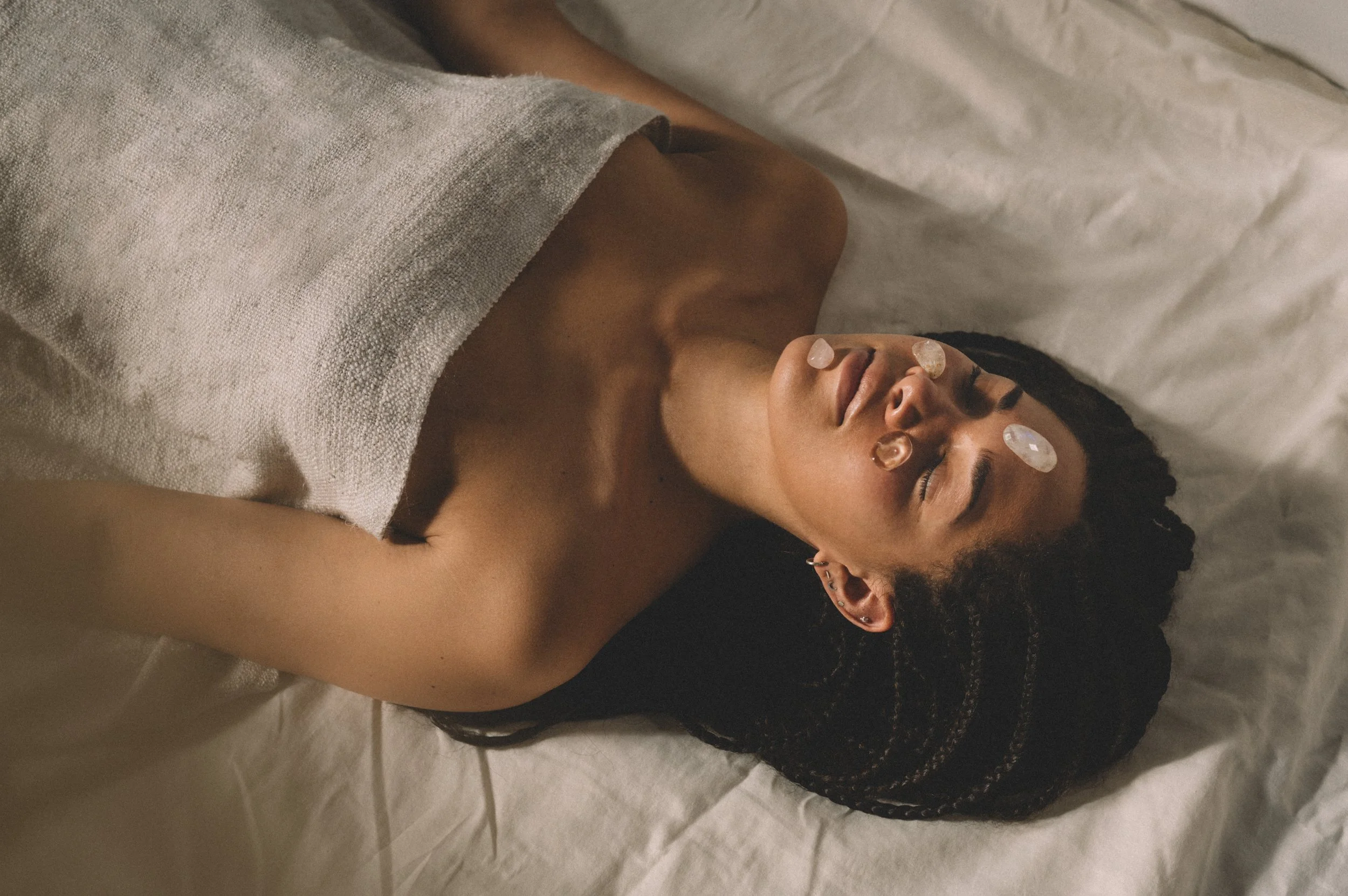 Person lying on a bed with crystals on their face, covered by a towel, in a relaxing spa setting.